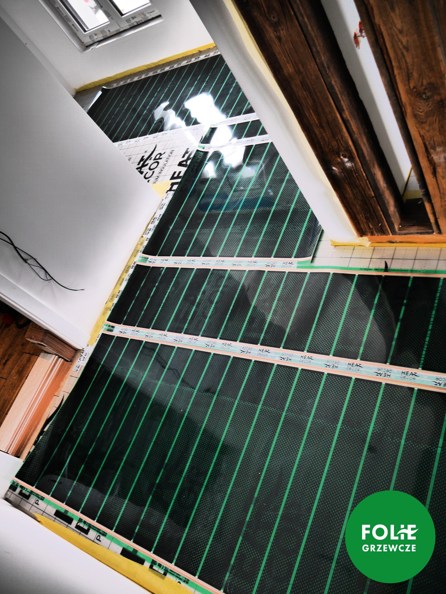 Rozłożone maty grzewcze z zielonymi liniami na podłodze w trakcie instalacji, widoczne elementy drewniane konstrukcji dachu i okno dachowe.