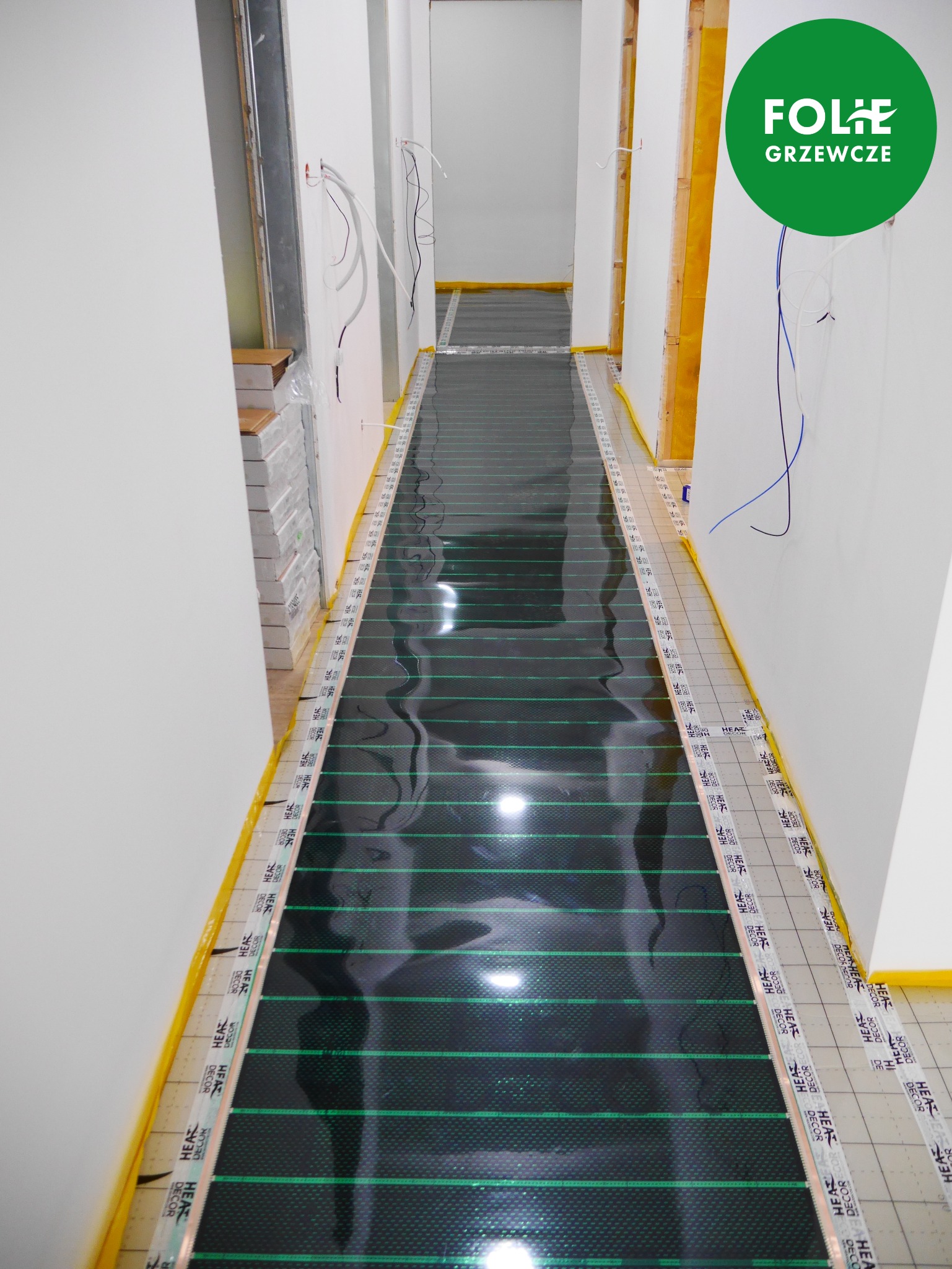 Instalacja folii grzewczej na podłodze w korytarzu, widoczne przewody elektryczne wystające ze ściany, taśmy mocujące z nadrukiem producenta wzdłuż krawędzi folii.
