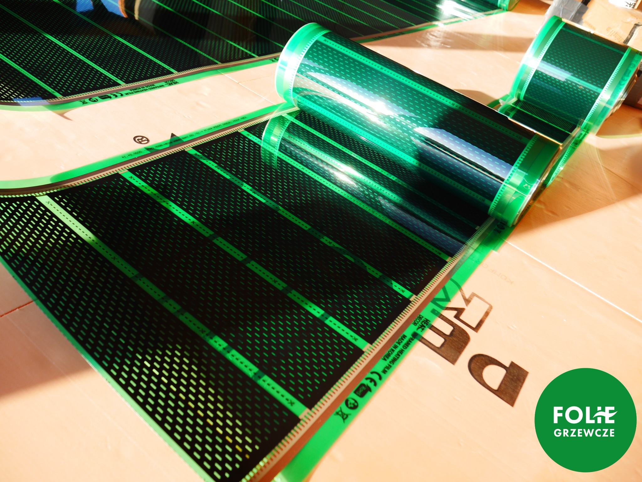 Rozwinięta i zwinięta w rolkę czarna folia grzewcza z zielonym obramowaniem na jasnej powierzchni, widoczne oznaczenia producenta i certyfikaty.