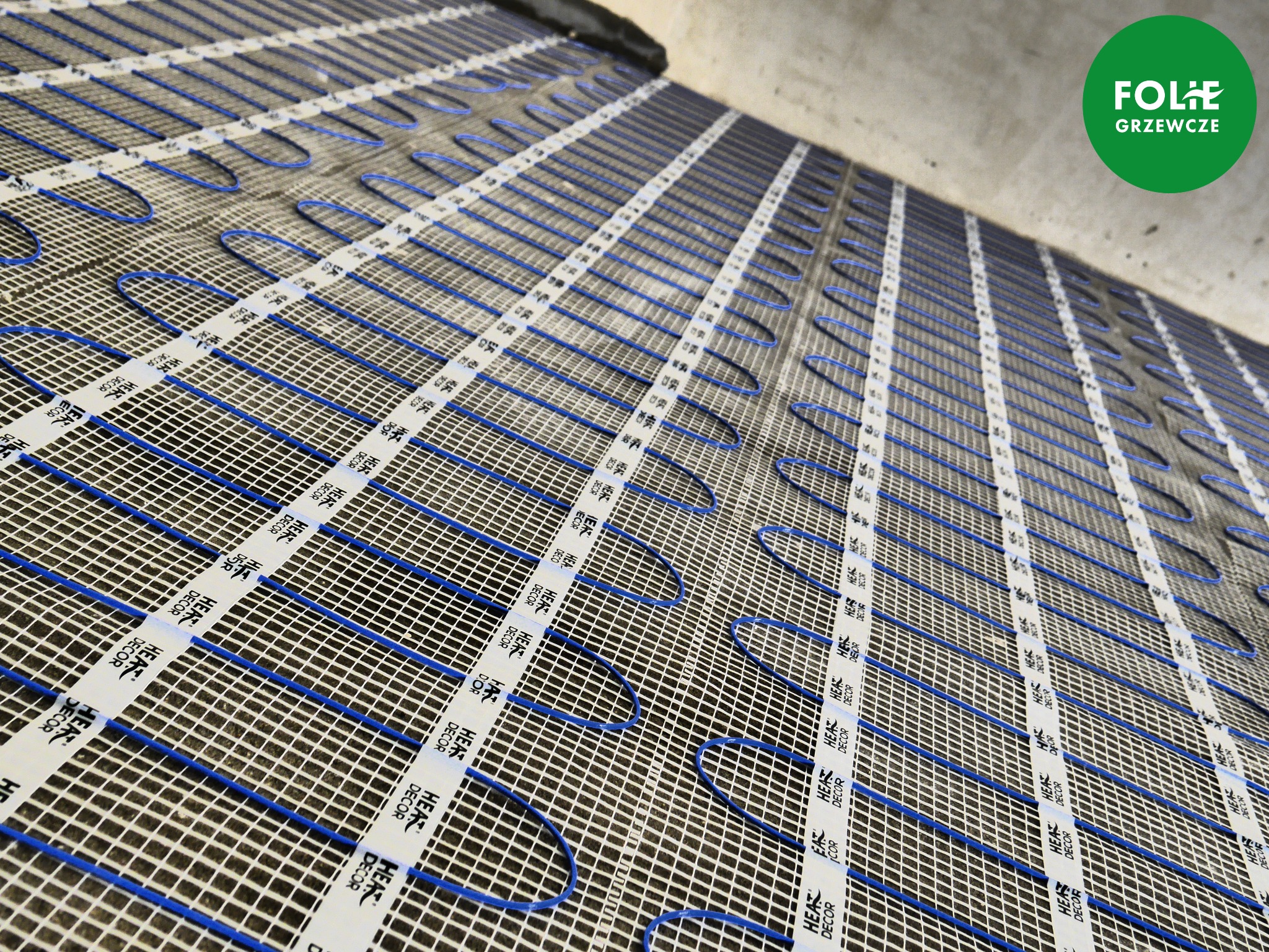 Niebieskie przewody grzewcze ułożone na siatce z białymi taśmami mocującymi z nadrukiem 'HEAT DECOR' przed wylewką, widoczne logo producenta w prawym górnym rogu.