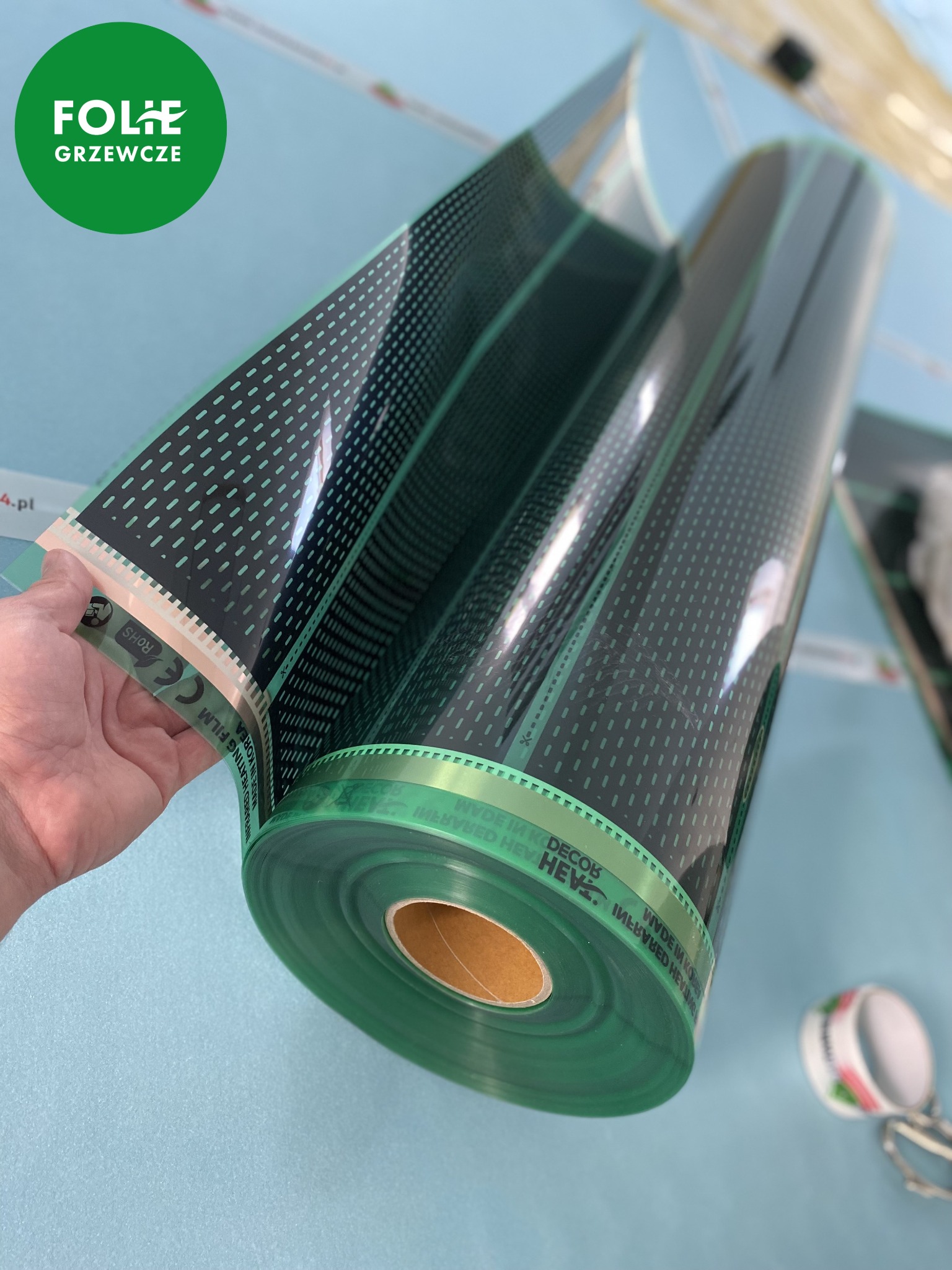 Rolka przezroczystej folii grzewczej z czarnym wzorem kropek, trzymana w dłoni, widoczne oznaczenia CE i RoHS na krawędzi folii.