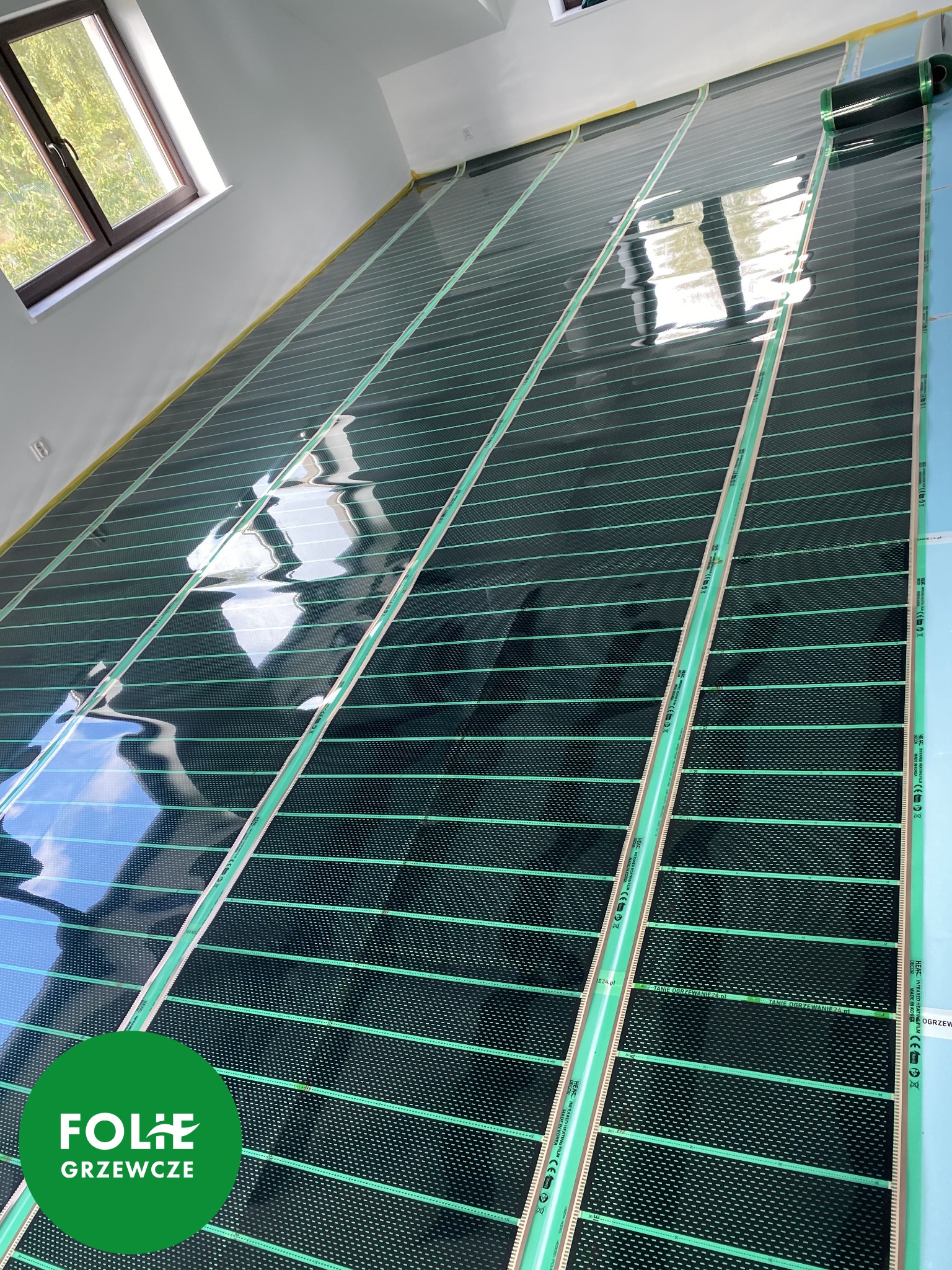 Montaż folii grzewczej na podłodze w nowym pomieszczeniu, widok z góry na rozłożone pasy folii z zielonymi paskami przewodzącymi, widoczne okno i fragment ściany.