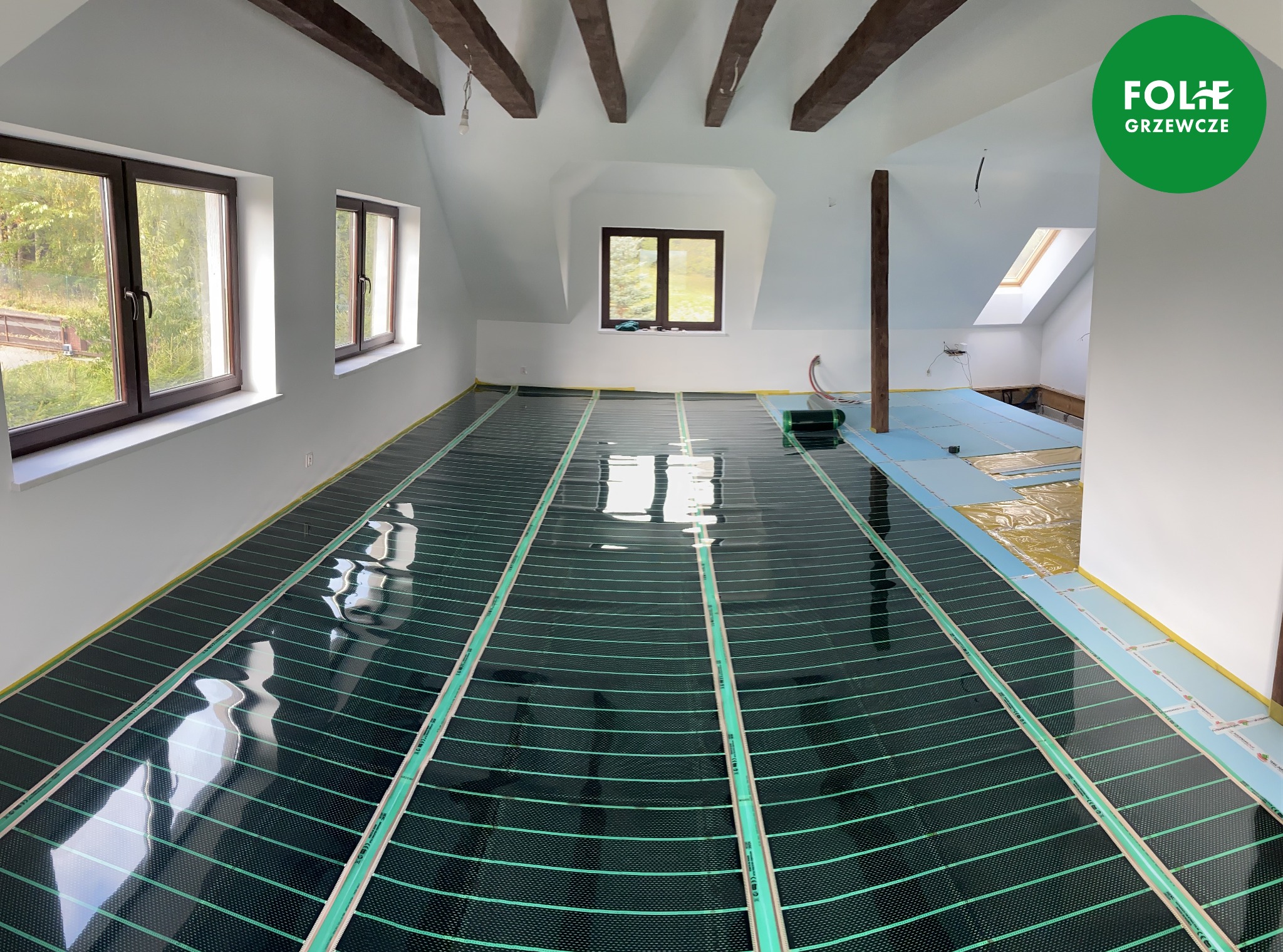 Montaż folii grzewczej na poddaszu z drewnianymi belkami, widoczne okna dachowe i logo producenta w prawym górnym rogu.