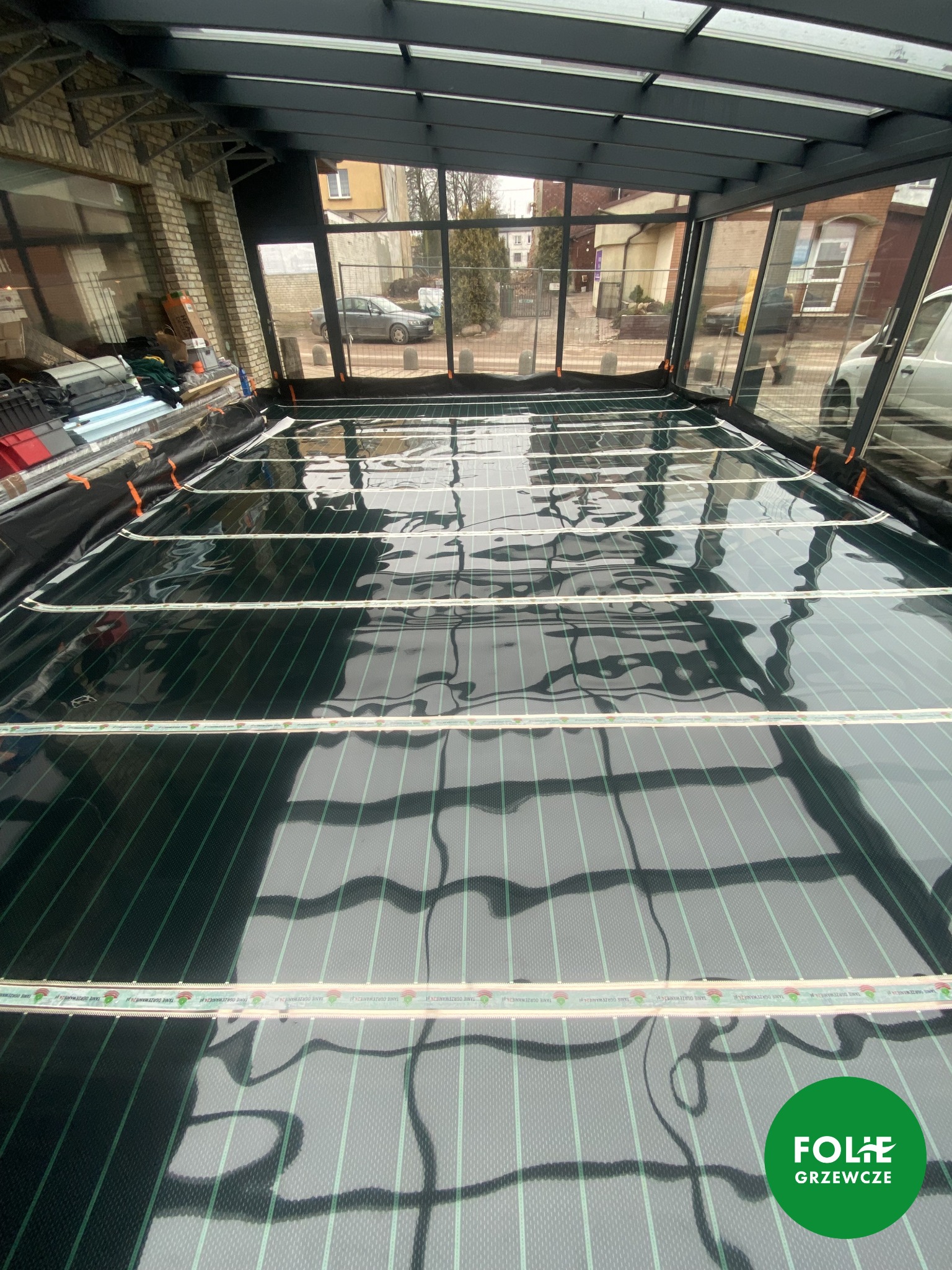Montaż folii grzewczej z widocznymi paskami zasilającymi, ułożonej na czarnej macie izolacyjnej wewnątrz przeszklonej konstrukcji, z widokiem na ulicę i budynki na zewnątrz.