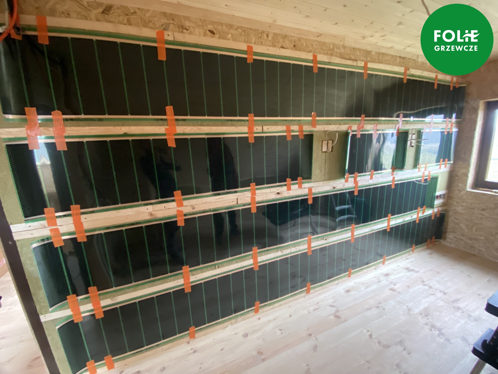 Montaż mat grzewczych na ścianie w drewnianym domu w Warszawie, folie grzewcze tymczasowo przytwierdzone pomarańczową taśmą.