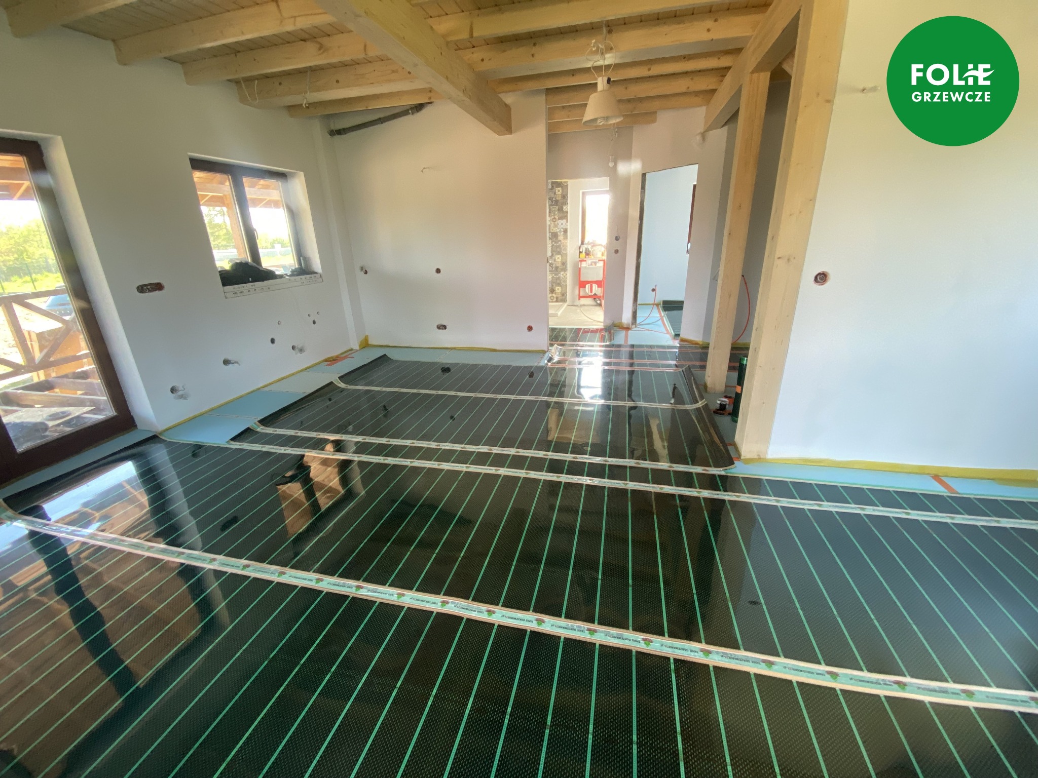 Montaż folii grzewczej na podłodze w surowym wnętrzu z drewnianym stropem i widocznym logo firmy instalującej w prawym górnym rogu.
