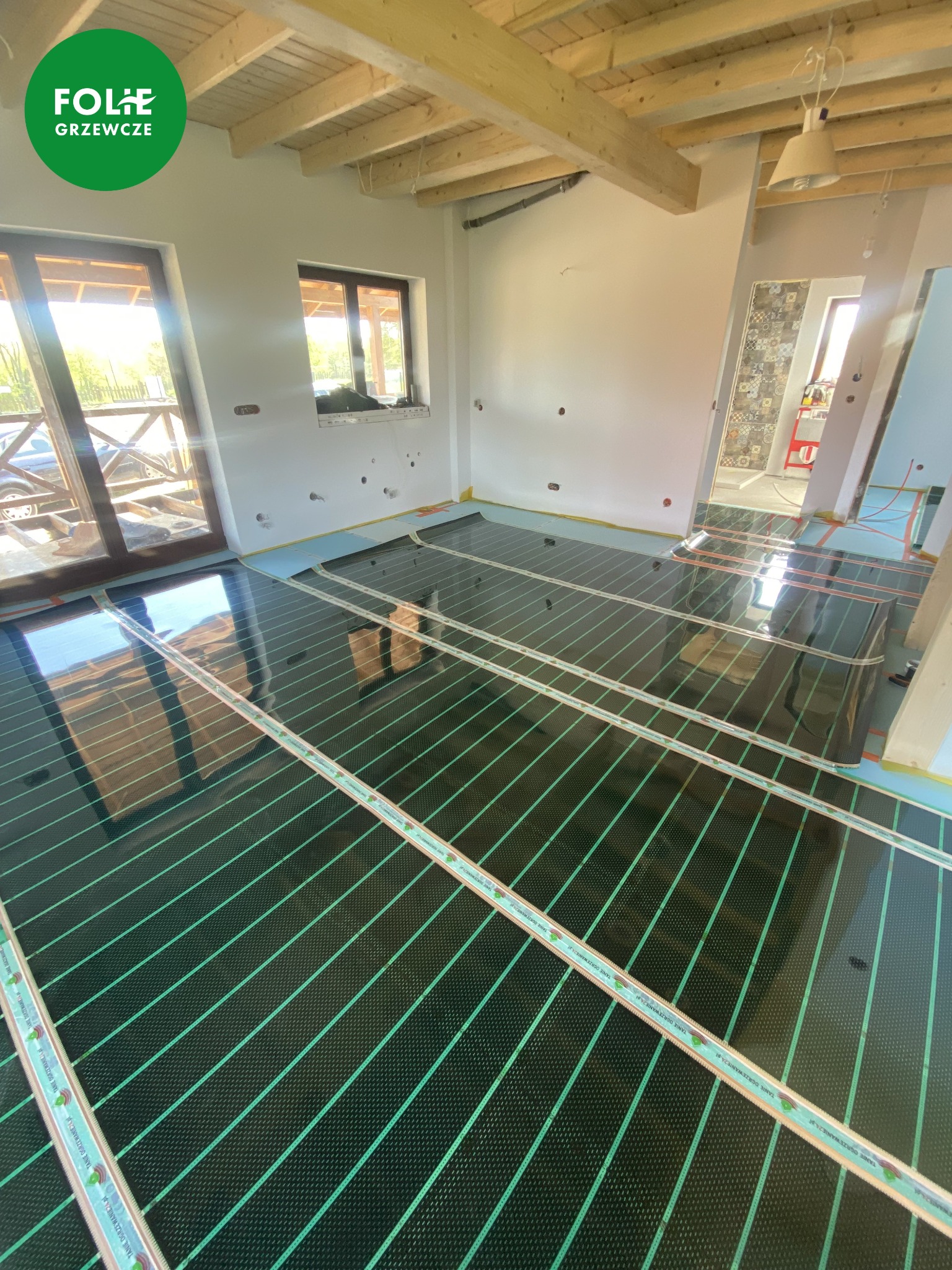 Montaż folii grzewczych na podłodze w jasnym pomieszczeniu z drewnianym stropem, widoczne okno z odbijającym się światłem słonecznym i logo producenta na folii.
