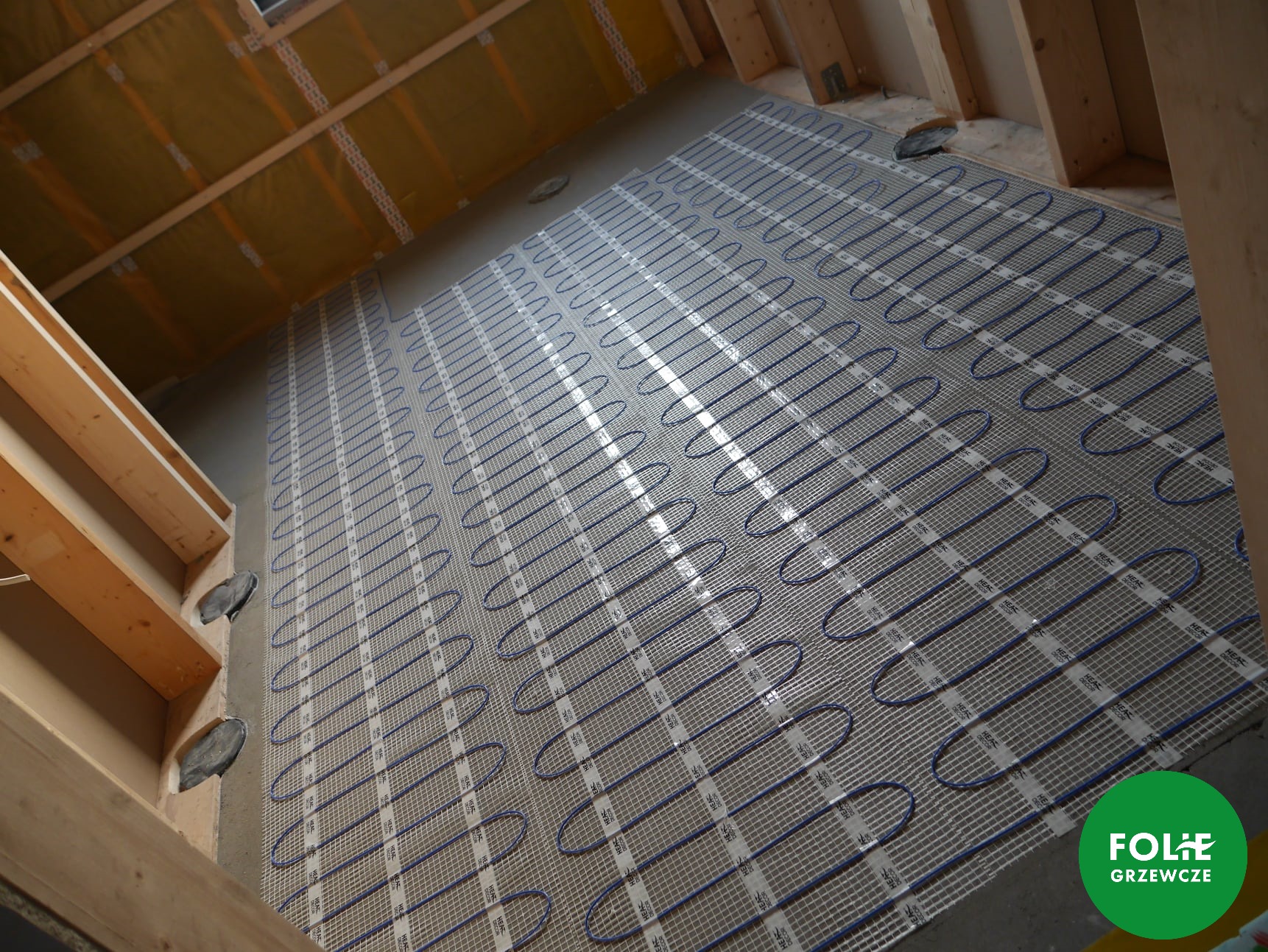 Rozłożona mata grzewcza z niebieskimi przewodami na siatce, przygotowana do zalania wylewką, widoczne drewniane elementy konstrukcji stropu i ściany, w rogu logo producenta.