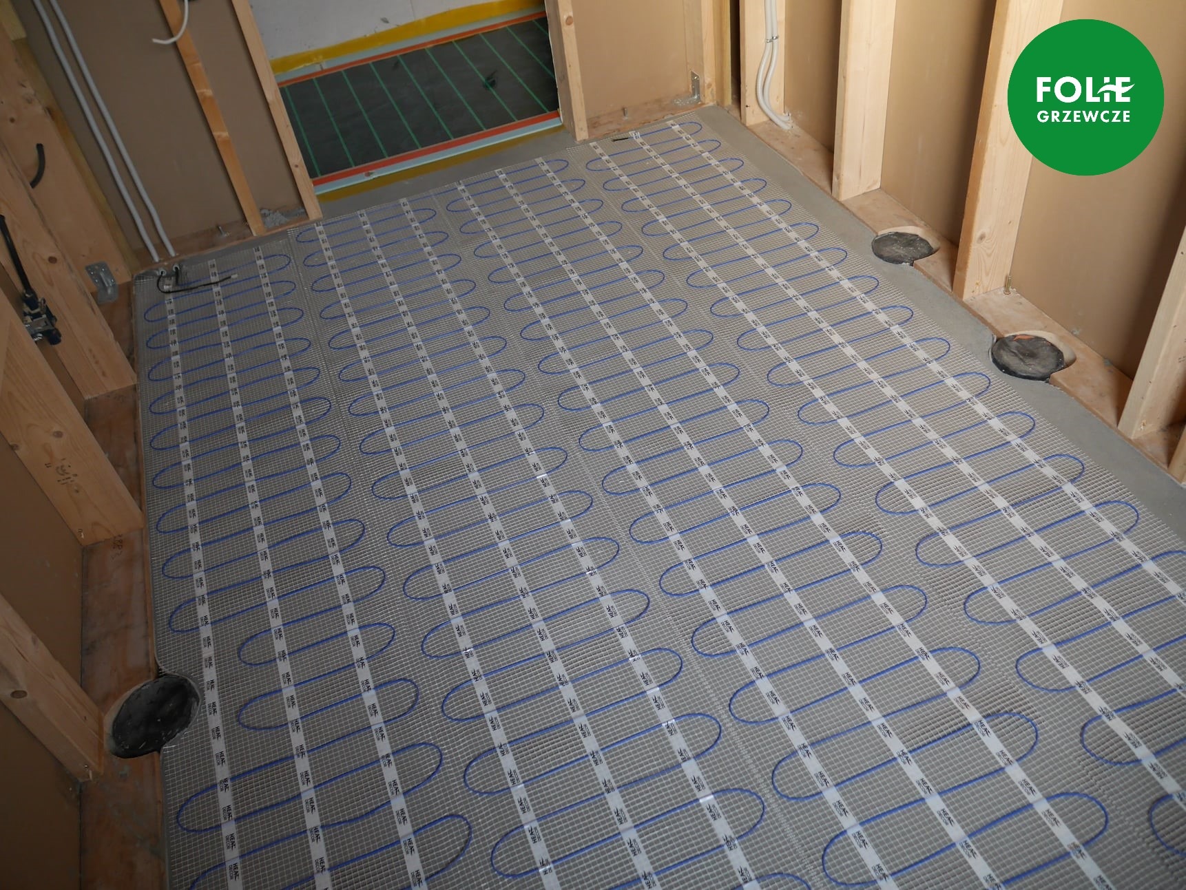 Szara mata z niebieskimi kablami grzewczymi ułożona na podłodze w trakcie montażu, widoczne drewniane elementy konstrukcyjne ścian i logo producenta w prawym górnym rogu.