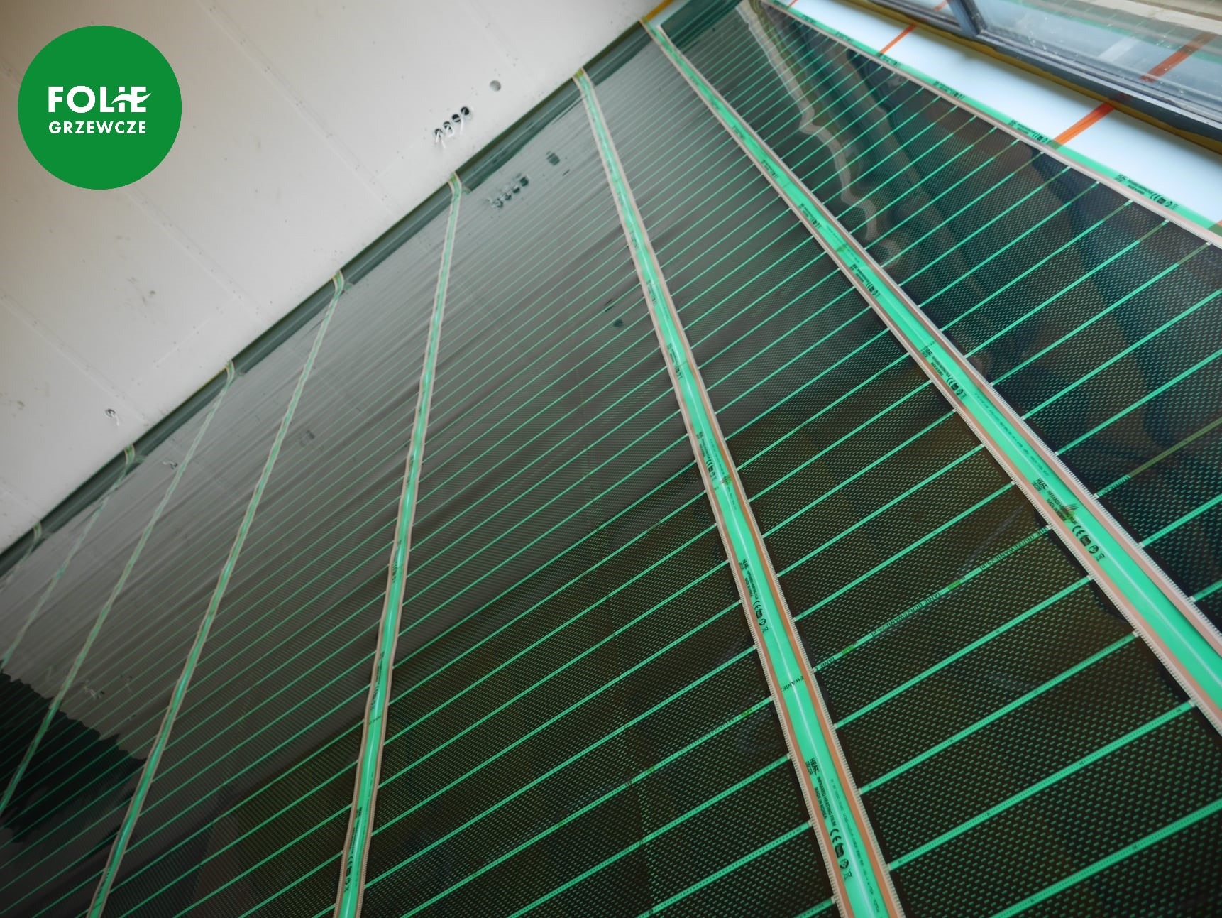 Rozłożona, czarna mata z zielonymi paskami na podłodze, przygotowana do montażu ogrzewania podłogowego, widoczny fragment ściany i okna.