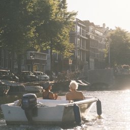 Nadya_Kova Photo - Dwie osoby płyną łodzią motorową po kanale w Amsterdamie, w tle kamienice i zieleń drzew w letnim słońcu.