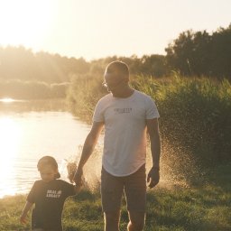 Nadya_Kova Photo - Ojciec w okularach i syn trzymają się za ręce, spacerując po trawie w promieniach zachodzącego słońca, z jeziorem i drzewami w tle.