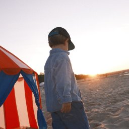 Nadya_Kova Photo - Chłopiec w dżinsowej koszuli i czapce z daszkiem idzie boso po piaszczystej plaży w kierunku zachodzącego słońca, obok stoi namiot plażowy w biało-czerwone pasy.
