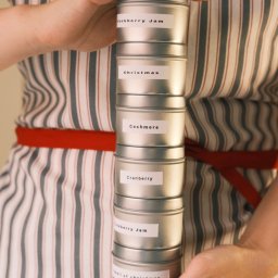 Nadya_Kova Photo - Osoba w fartuchu w paski trzyma stos srebrnych puszek z etykietami: Blackberry Jam, Christmas, Cashmere, Cranberry, Blackberry Jam, Smell of Christmas.