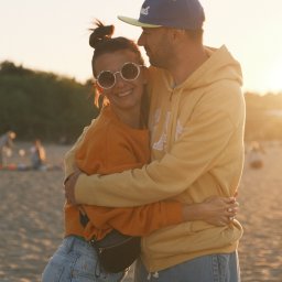 Nadya_Kova Photo - Uśmiechnięta para w objęciach na plaży o zachodzie słońca, kobieta w pomarańczowej bluzie i okularach w stylu retro, mężczyzna w żółtej bluzie z kapturem i czapce z daszkiem.