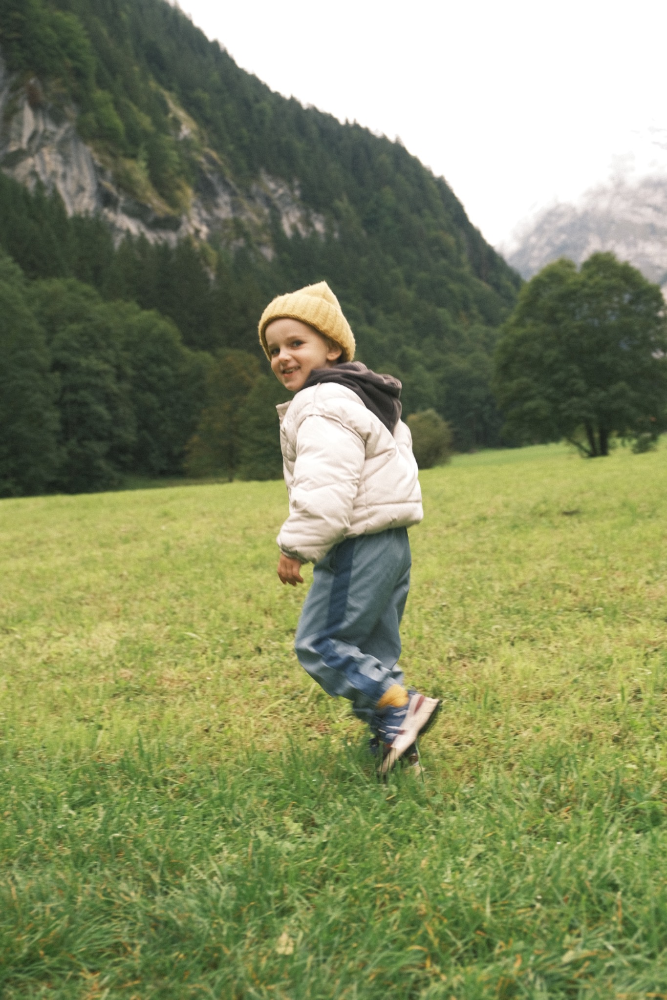 Chłopiec w żółtej czapce i puchowej kurtce biegnie po zielonej łące z widokiem na zalesione góry.