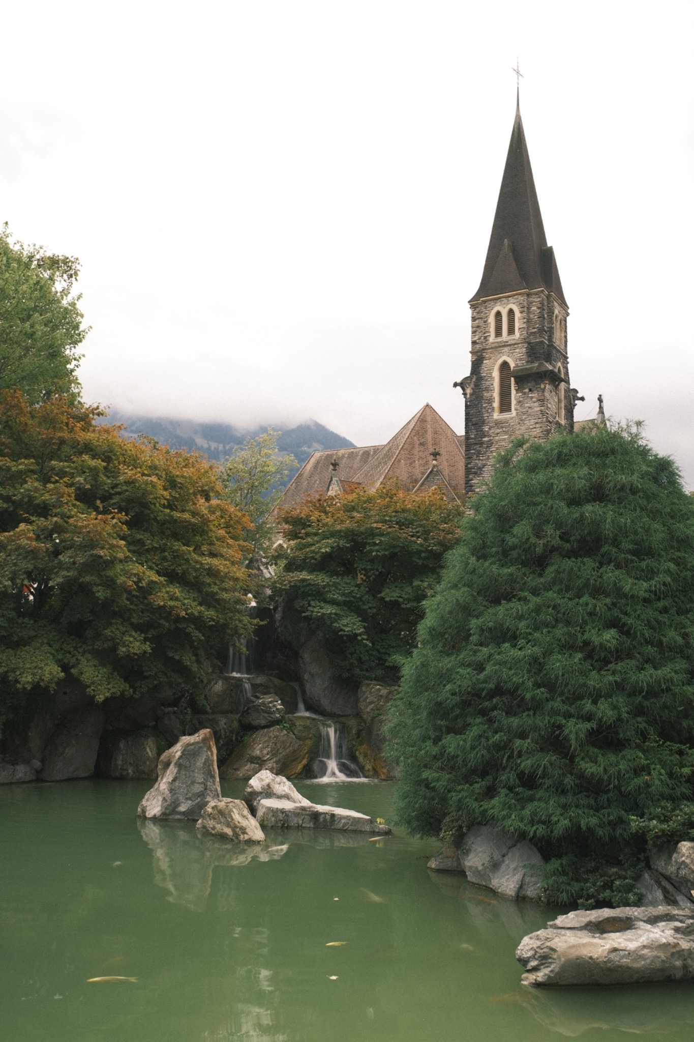 Kamienny kościół z wieżą i spiczastym dachem, otoczony zielenią drzew i kaskadami wodospadu w tle gór pokrytych mgłą.