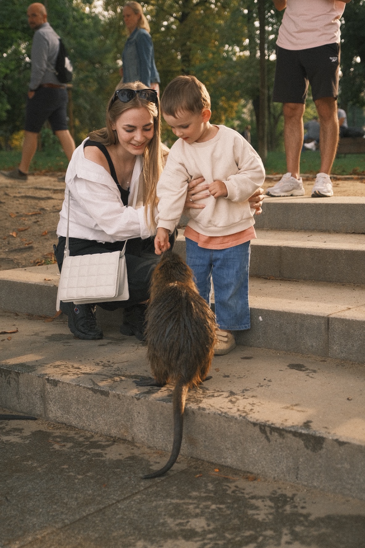 Kobieta z dzieckiem karmią nutrie na betonowych schodach w parku, w tle spacerujący ludzie.