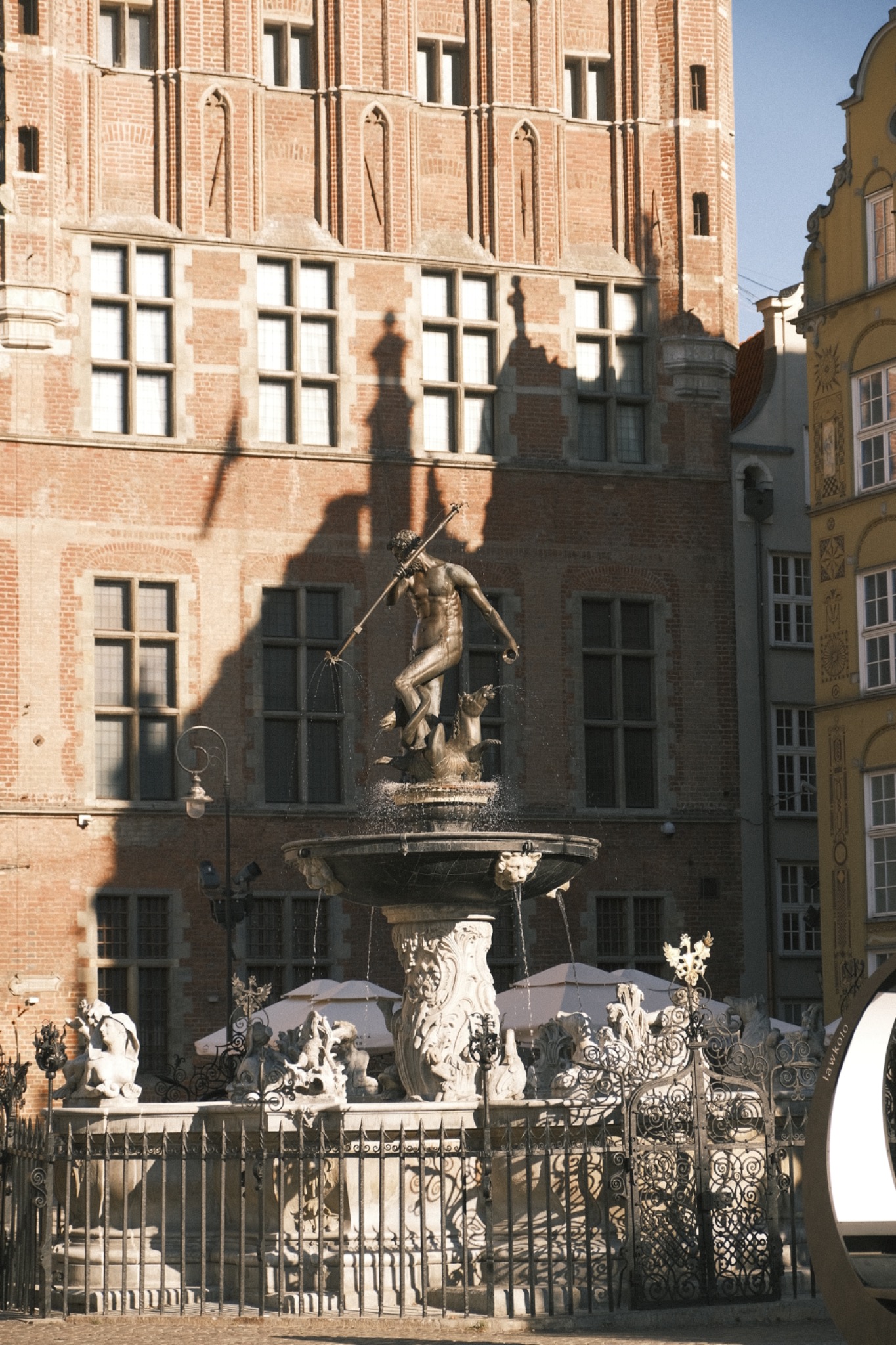 Fontanna Neptuna w Gdańsku z widocznym cieniem rzucanym na ceglaną fasadę budynku w tle.