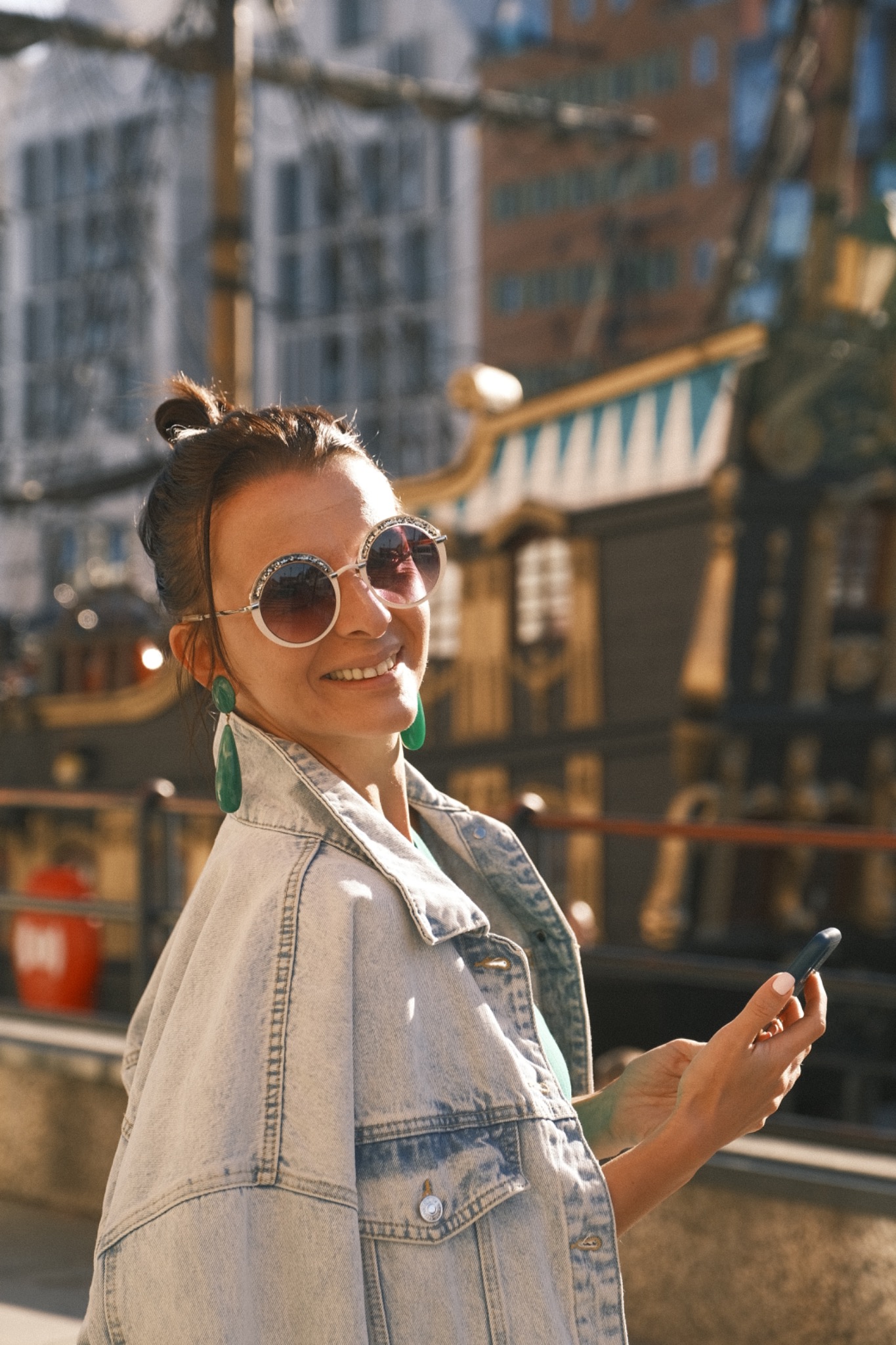 Uśmiechnięta kobieta w okularach i dżinsowej kurtce, trzymająca telefon na tle budynku w stylu statku.