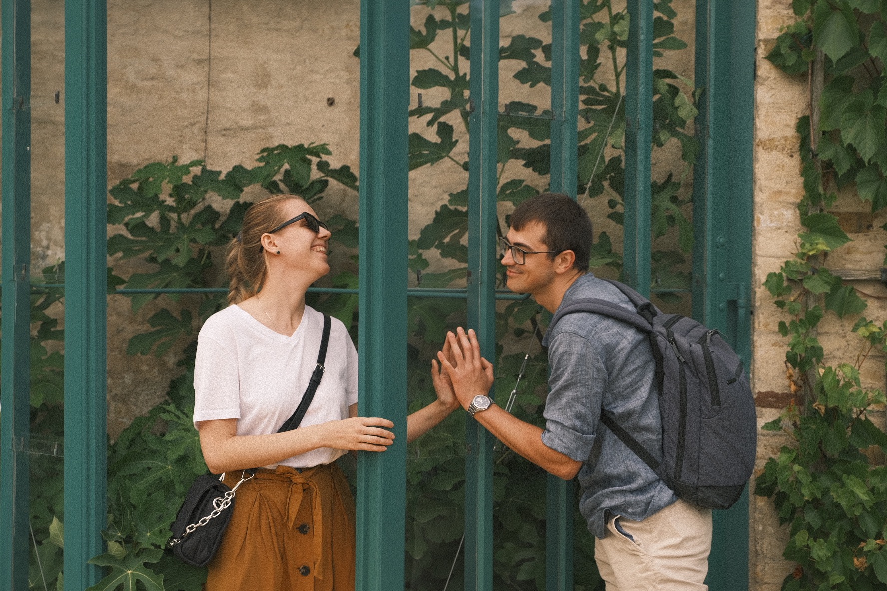 Uśmiechnięta para, dziewczyna w okularach przeciwsłonecznych i chłopak z plecakiem, dotykają dłońmi przez zieloną, metalową kratę porośniętą bluszczem.