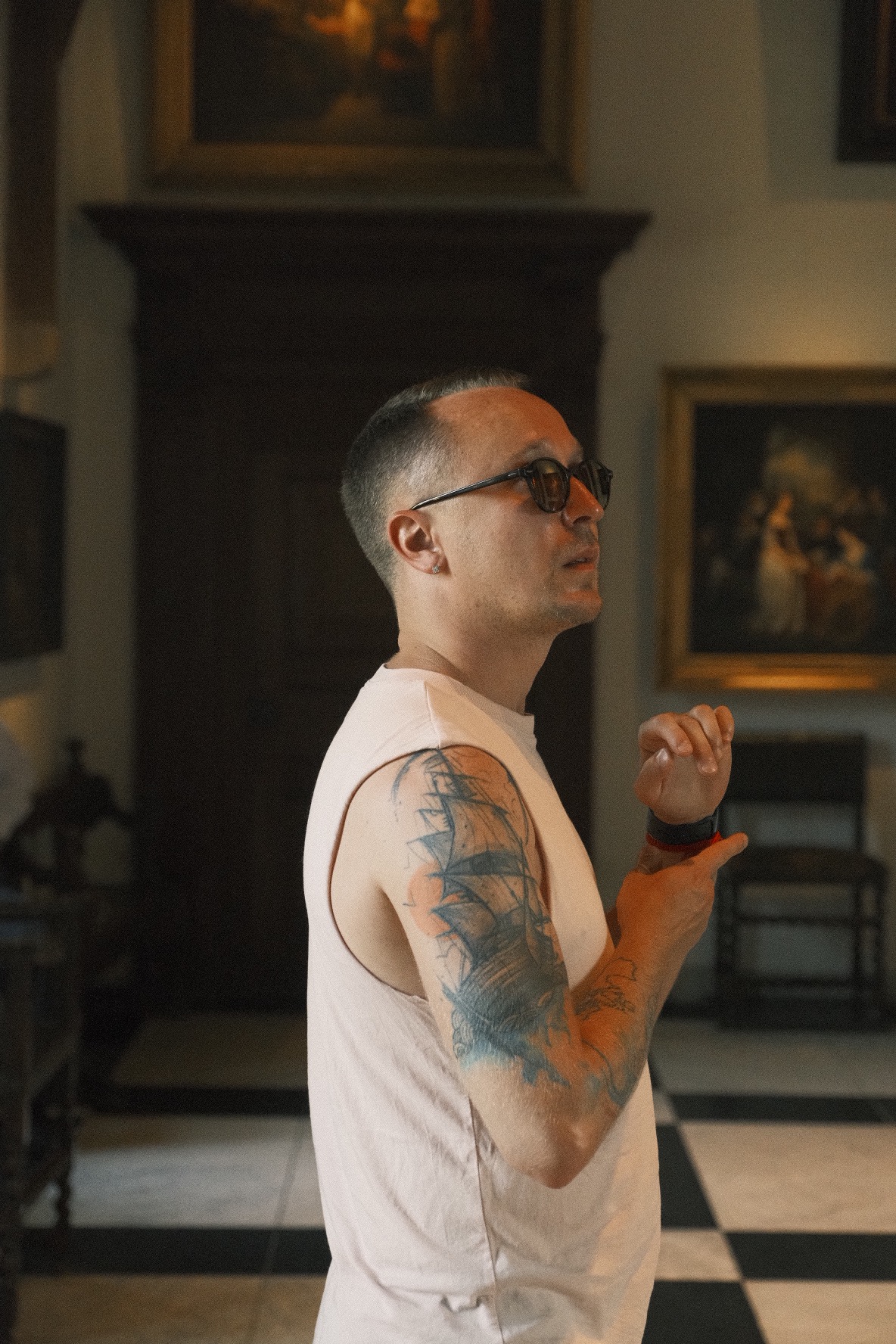 Mężczyzna w okularach i podkoszulku bez rękawów z tatuażem żaglowca na ramieniu, stojący w eleganckim wnętrzu z obrazami i czarno-białą podłogą.