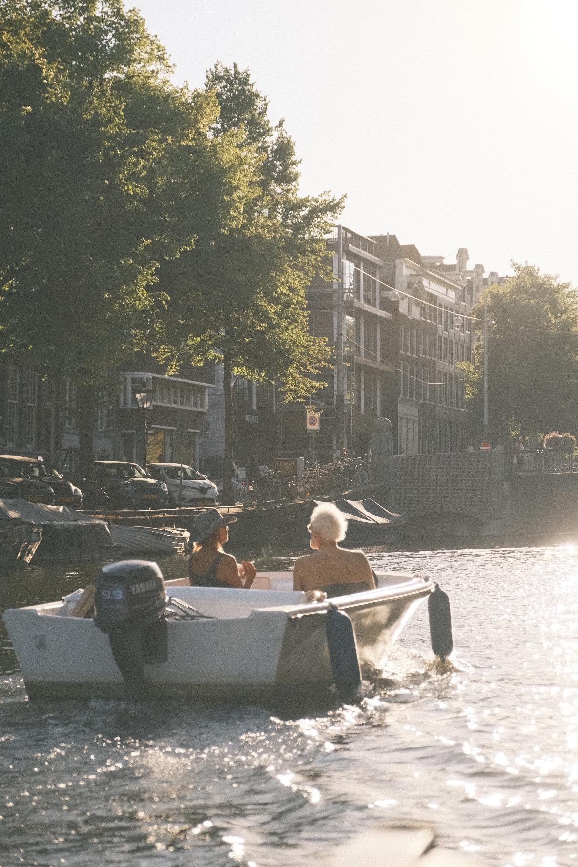 Dwie osoby płyną łodzią motorową po kanale w Amsterdamie, w tle kamienice i zieleń drzew w letnim słońcu.