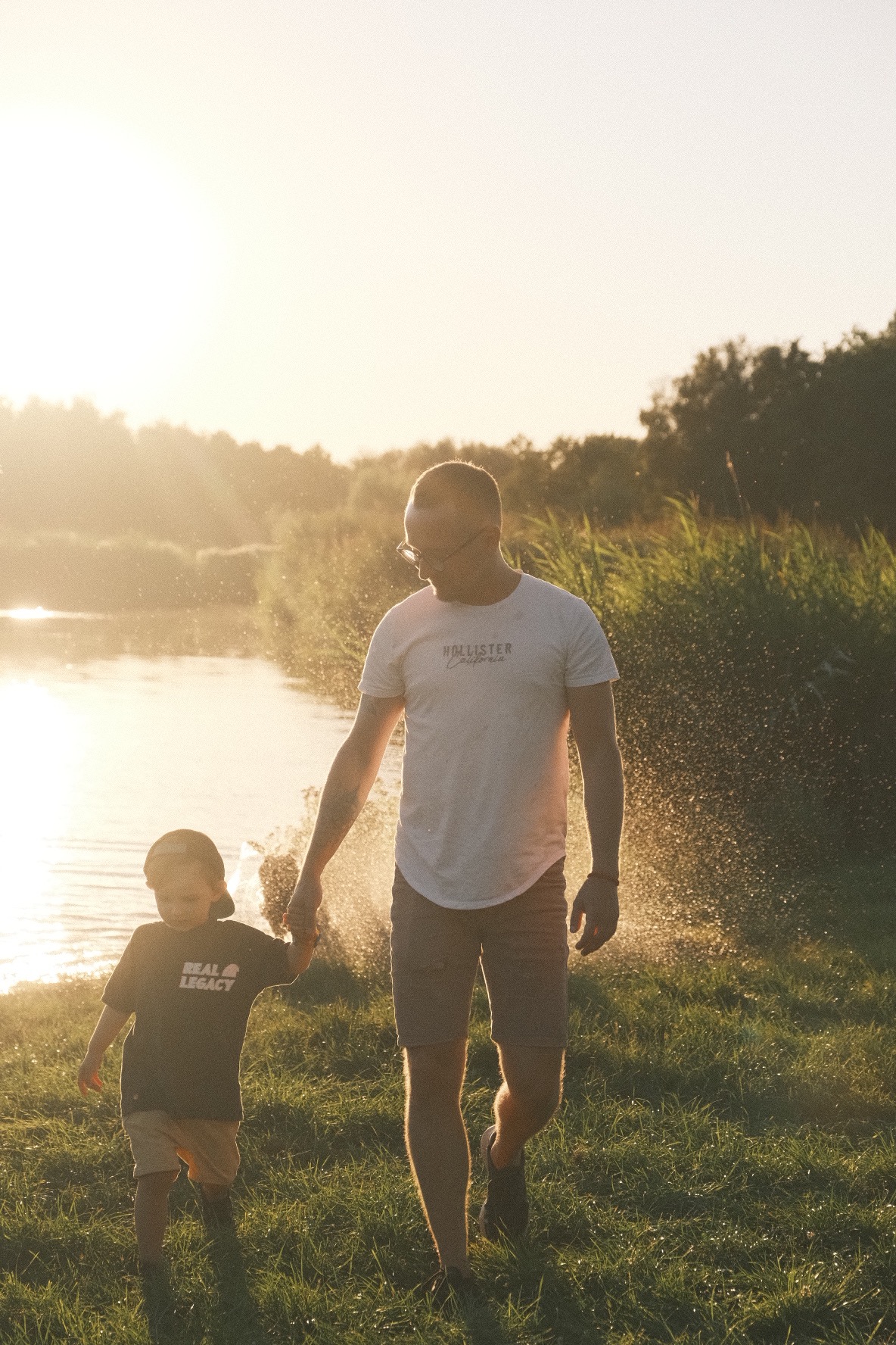 Ojciec w okularach i syn trzymają się za ręce, spacerując po trawie w promieniach zachodzącego słońca, z jeziorem i drzewami w tle.
