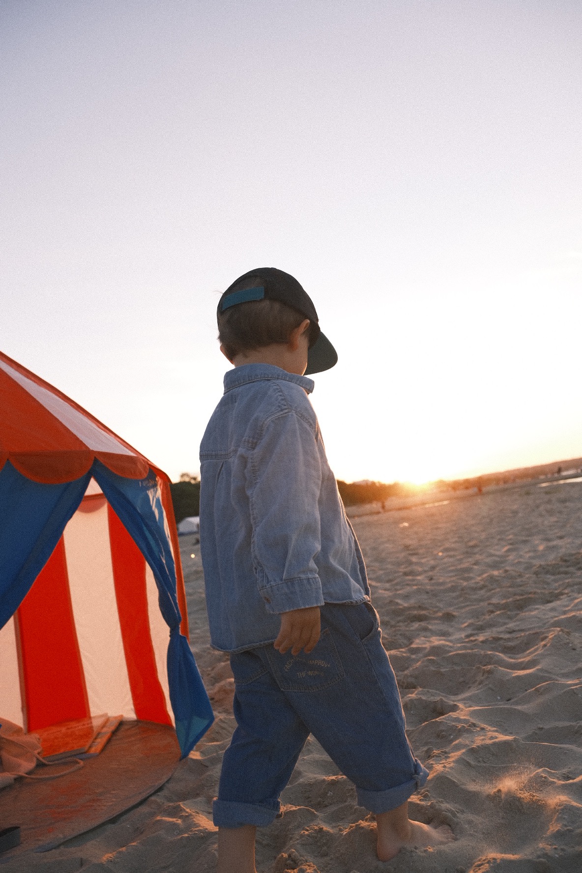 Chłopiec w dżinsowej koszuli i czapce z daszkiem idzie boso po piaszczystej plaży w kierunku zachodzącego słońca, obok stoi namiot plażowy w biało-czerwone pasy.