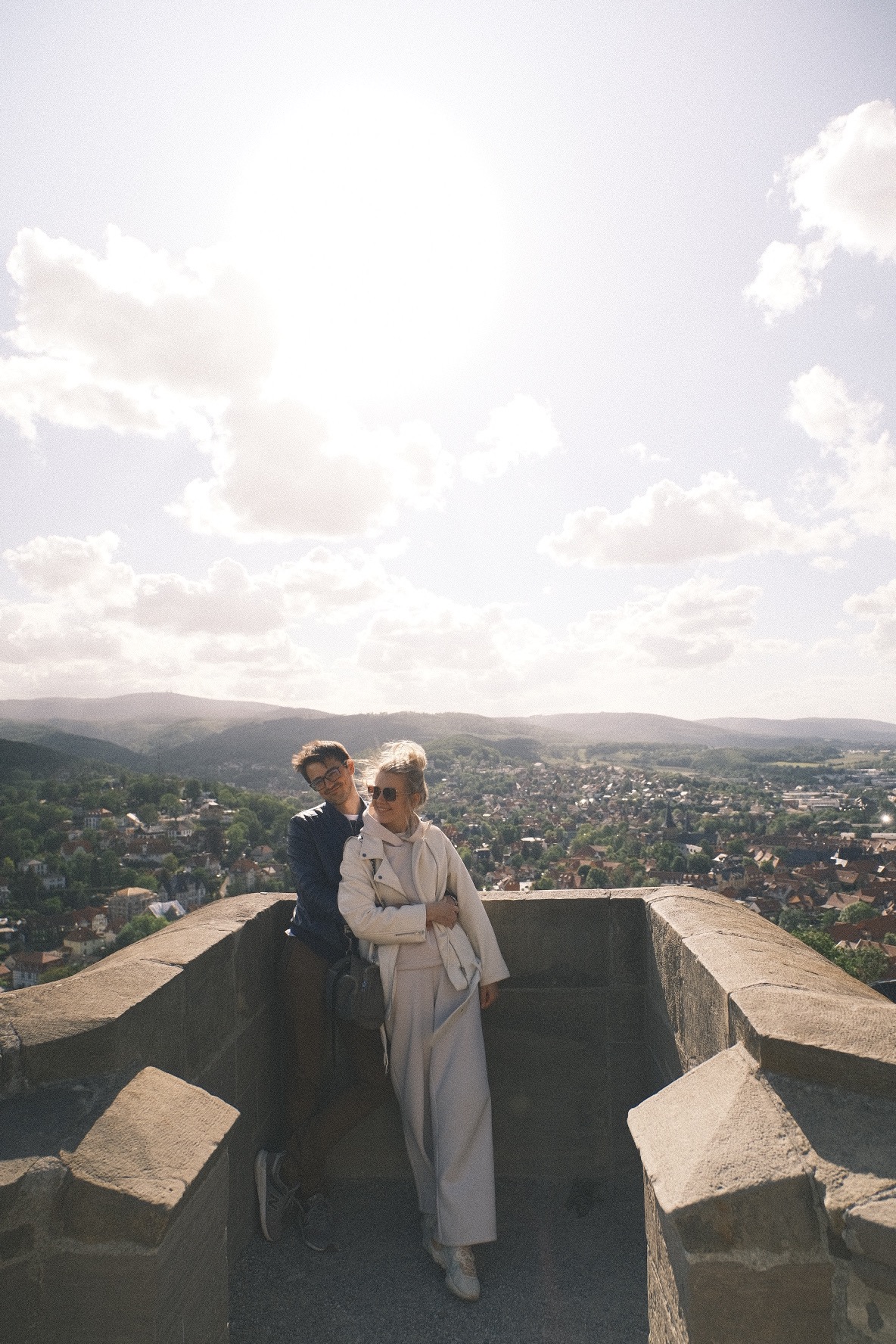 Para w jasnych ubraniach na tle panoramy miasta i wzgórz, zrobione z punktu widokowego na kamiennej konstrukcji w słoneczny dzień.