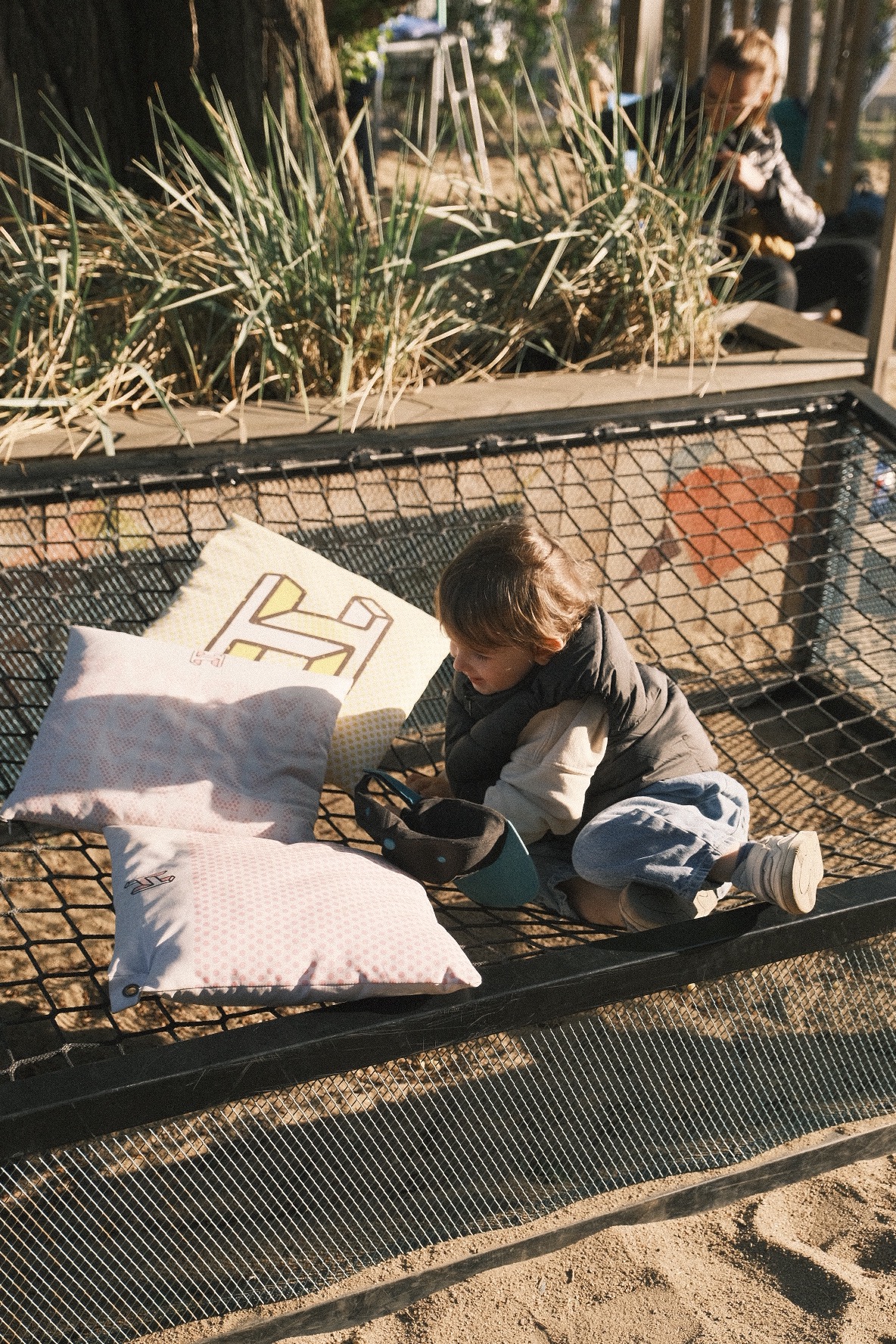 Chłopiec siedzi na hamaku z siatki, otoczony poduszkami, w tle widoczna osoba siedząca na ławce i roślinność.
