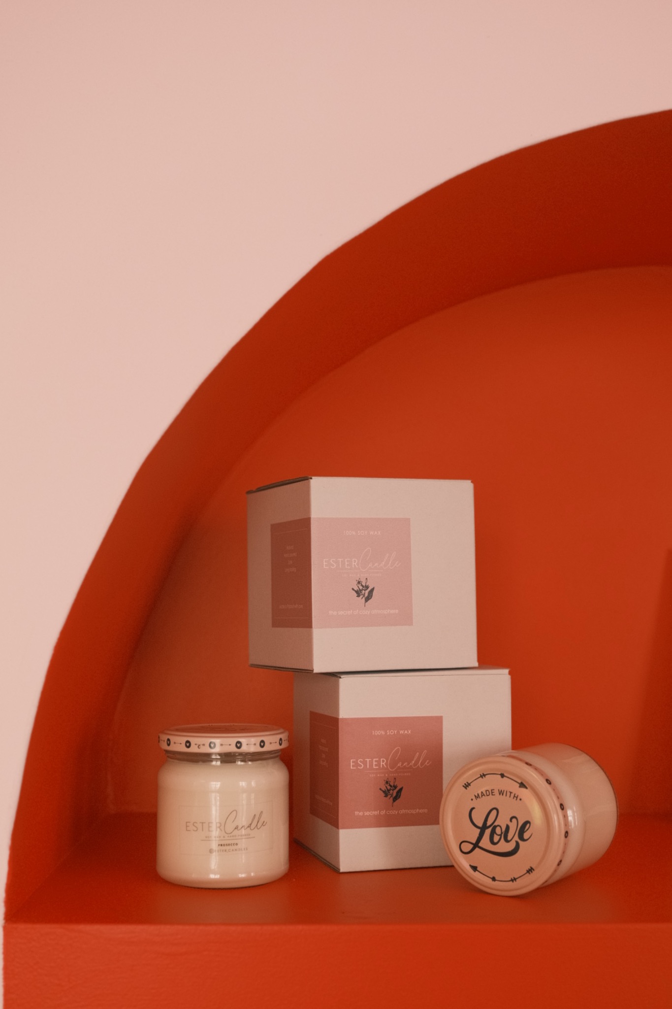 Ręcznie robione świece sojowe marki Ester Candle w minimalistycznych opakowaniach, ustawione na półce w niszy o pomarańczowym kolorze.