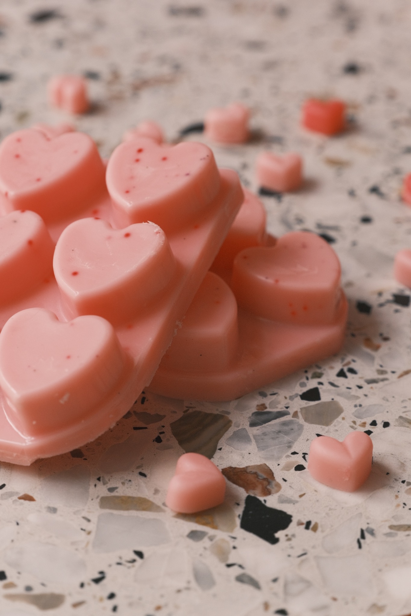 Różowe woski zapachowe w kształcie serc z delikatnymi czerwonymi plamkami, ułożone na marmurowej powierzchni z rozsypanymi pojedynczymi serduszkami.