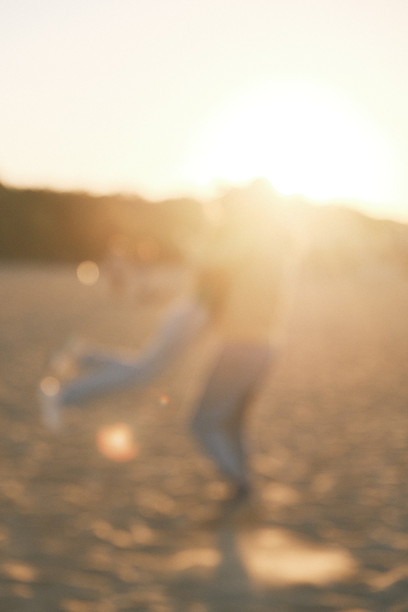 Rozmazane sylwetki pary na plaży o zachodzie słońca, kobieta unoszona w górę, ciepłe, rozproszone światło.