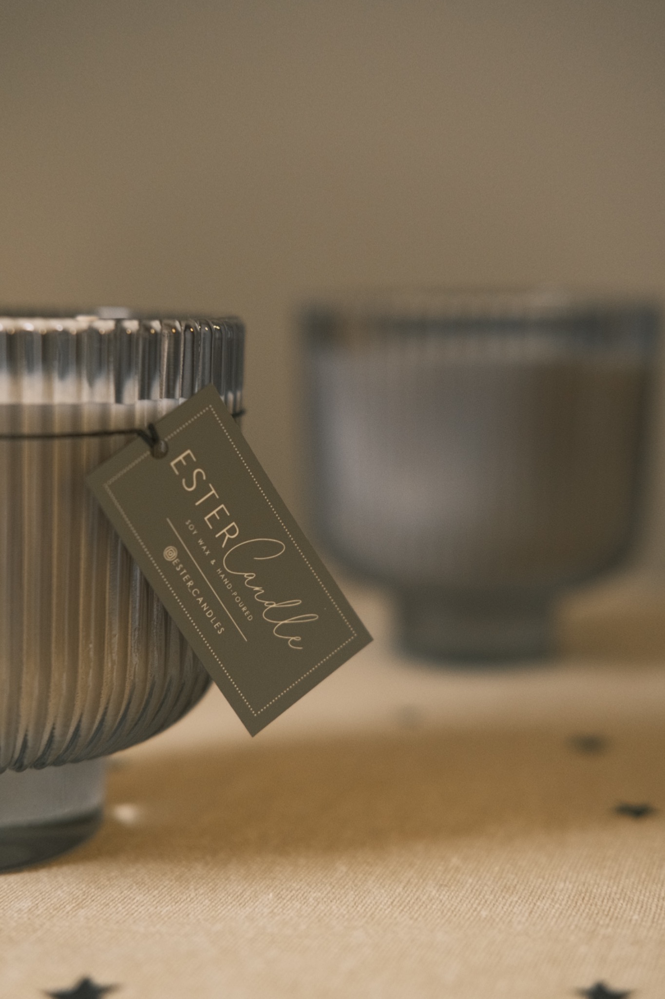 Szara świeca sojowa Ester Candle z prążkowaną fakturą szkła i etykietą, na beżowym obrusie z czarnymi gwiazdkami, druga świeca w tle.