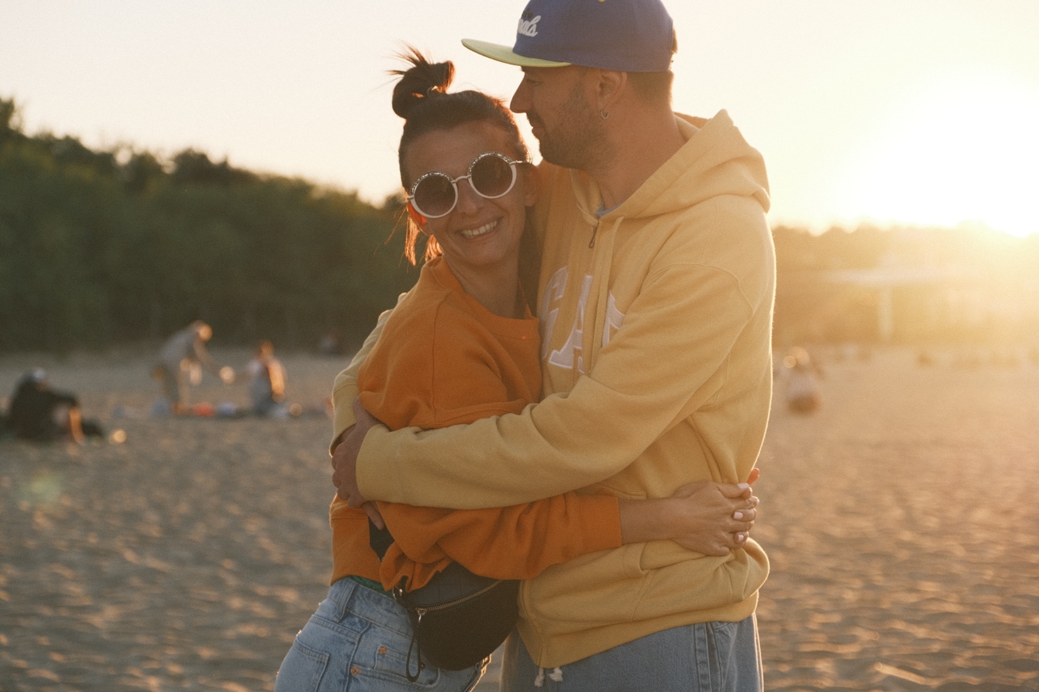 Uśmiechnięta para w objęciach na plaży o zachodzie słońca, kobieta w pomarańczowej bluzie i okularach w stylu retro, mężczyzna w żółtej bluzie z kapturem i czapce z daszkiem.
