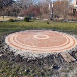 Sal-Bruk Dominik Salamon - Okrągły plac z kostki brukowej w odcieniach beżu i brązu, otoczony ziemią i trawą, z widoczną deską i młodymi drzewkami w tle.