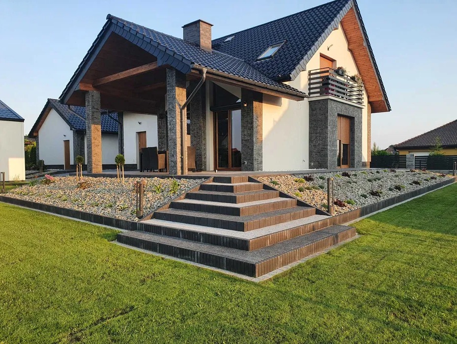 Nowoczesny dom jednorodzinny z tarasem wyłożonym kostką brukową, ozdobnym ogrodem skalnym i oświetleniem zewnętrznym, widok z poziomu trawnika.