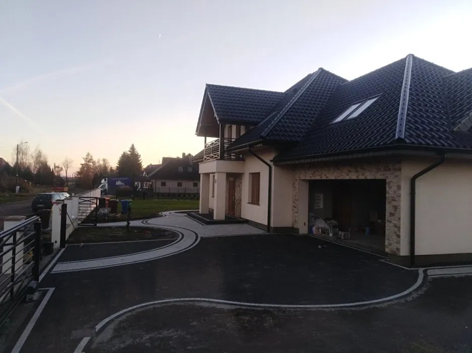 Nowoczesny dom jednorodzinny z czarnym dachem i jasną elewacją, z nowo ułożoną, ciemną kostką brukową z białymi obrzeżami, tworzącą faliste wzory na podjeździe.