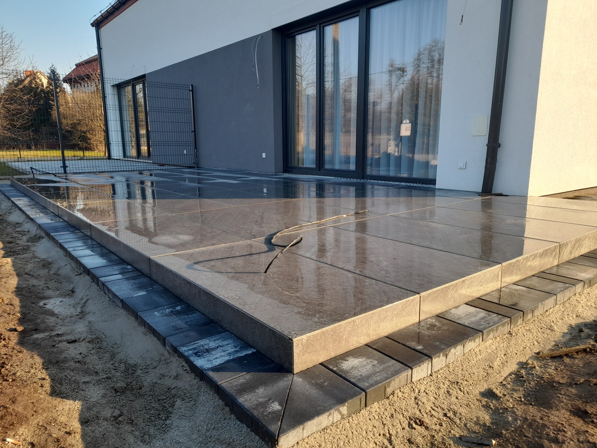 Nowoczesny taras z szarych, dużych płyt, obramowany ciemniejszą kostką, z widocznymi refleksami wody na powierzchni, przylegający do ściany budynku z dużymi oknami.
