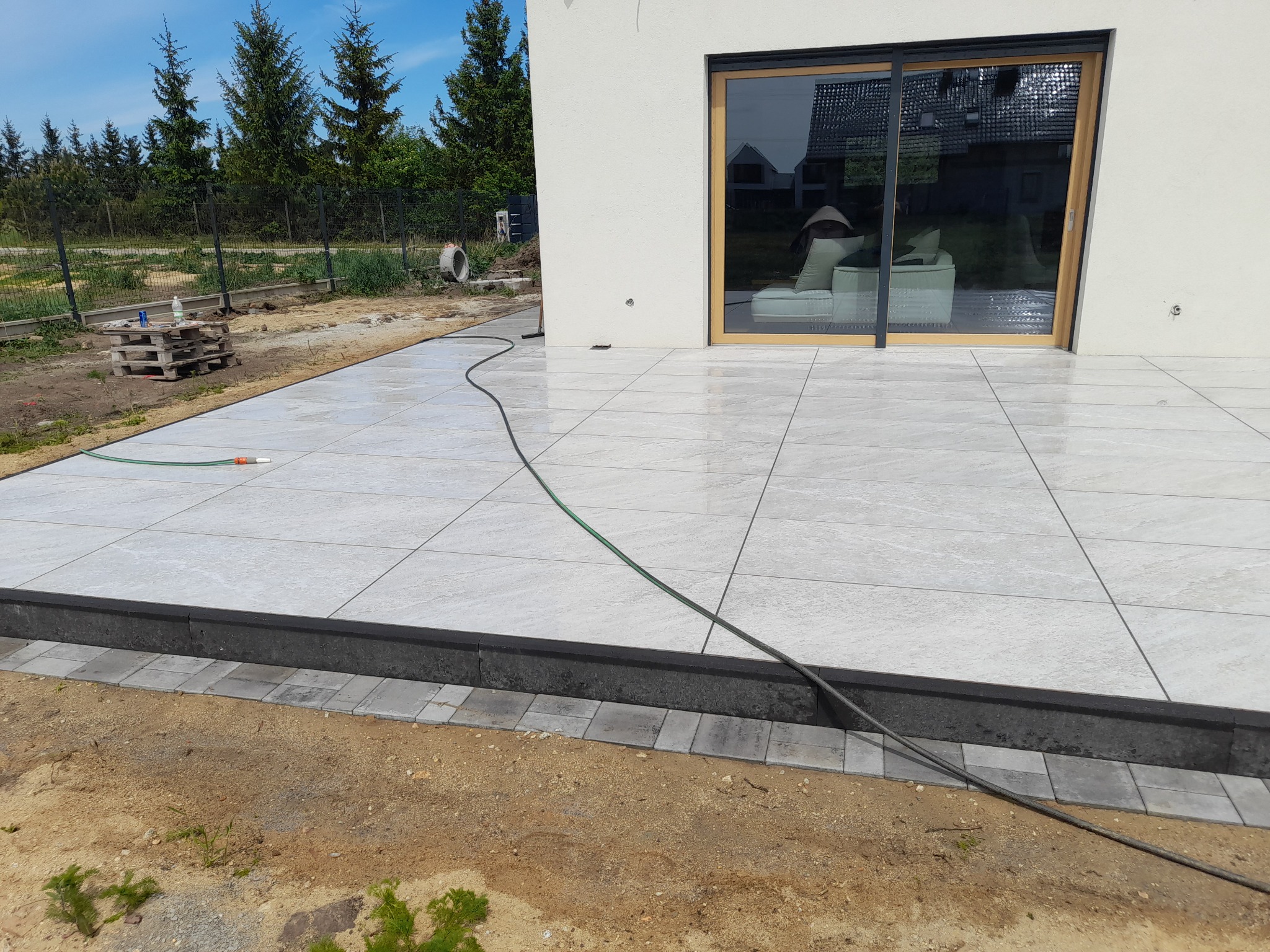 Jasnoszare, duże płyty tarasowe ułożone przed domem, z widoczną obrzeżem z kostki brukowej i czarnym krawężnikiem. W tle sosny i okno tarasowe.