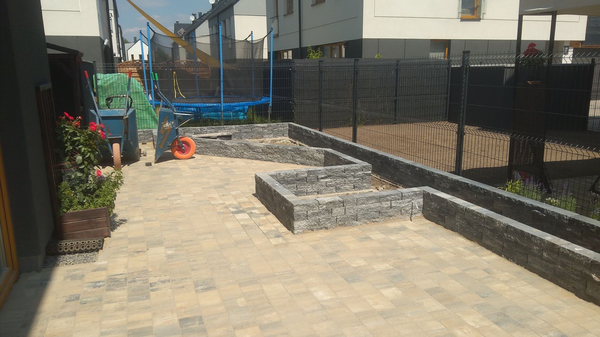 Ułożona kostka brukowa w odcieniach beżu i szarości tworząca patio z podwyższonymi rabatami z szarego kamienia, w tle siatka ogrodzeniowa i trampolina.