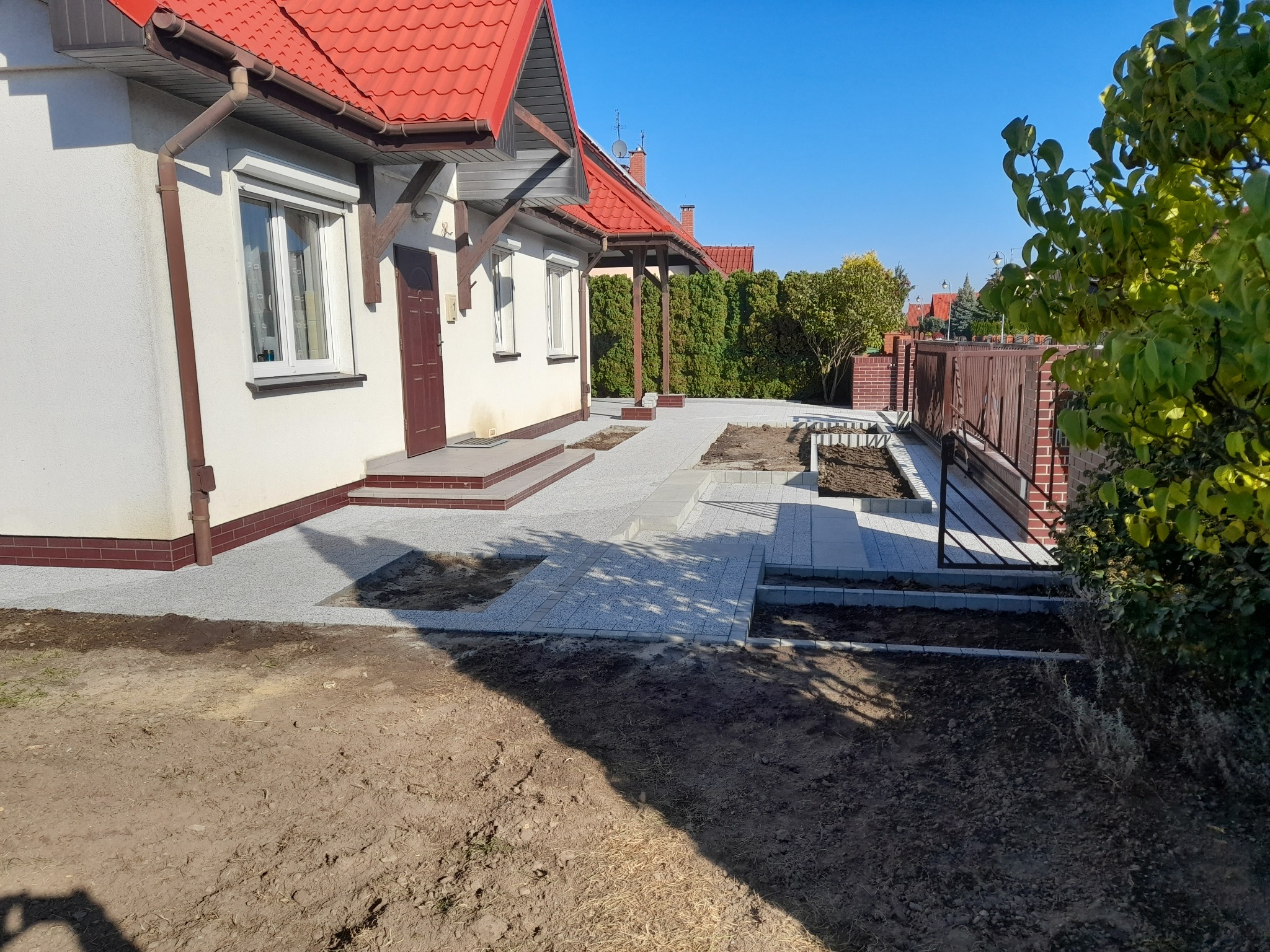 Nowo ułożony, jasnoszary chodnik z kostki brukowej prowadzący do domu z czerwoną dachówką, widoczne przygotowane rabaty kwiatowe i elementy ogrodzenia.