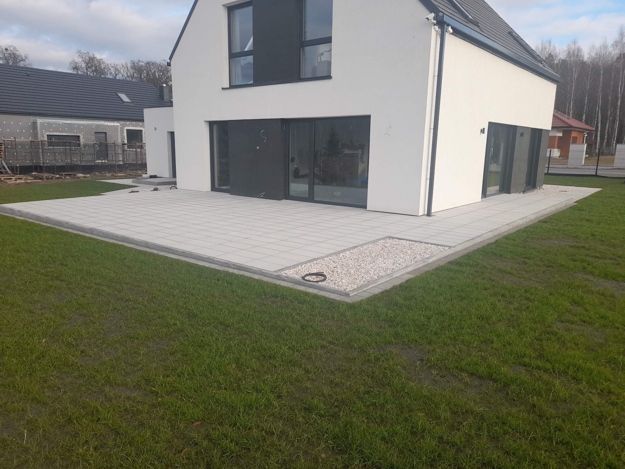 Nowoczesny taras z jasnoszarej kostki brukowej przy białym domu z dużymi przesuwnymi oknami, obrzeże z jasnych kamyków i czarnym wężem ogrodowym, trawnik z widocznymi śladami po deszczu.