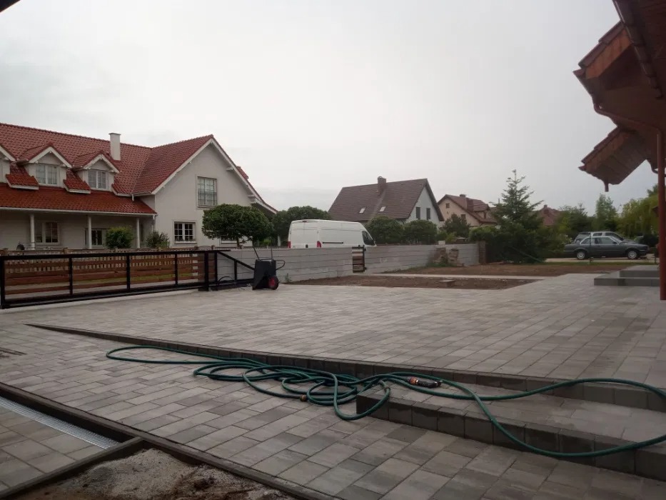 Ułożona szara kostka brukowa na podjeździe przed domem, widoczny wąż ogrodowy, brama wjazdowa i zaparkowany biały van w tle.
