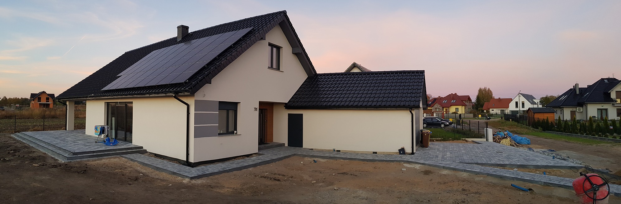 Nowoczesny dom jednorodzinny z panelami słonecznymi na dachu, świeżo ułożonym chodnikiem z szarej kostki brukowej i przygotowanym terenem pod trawnik, fotografowany o zmierzchu.