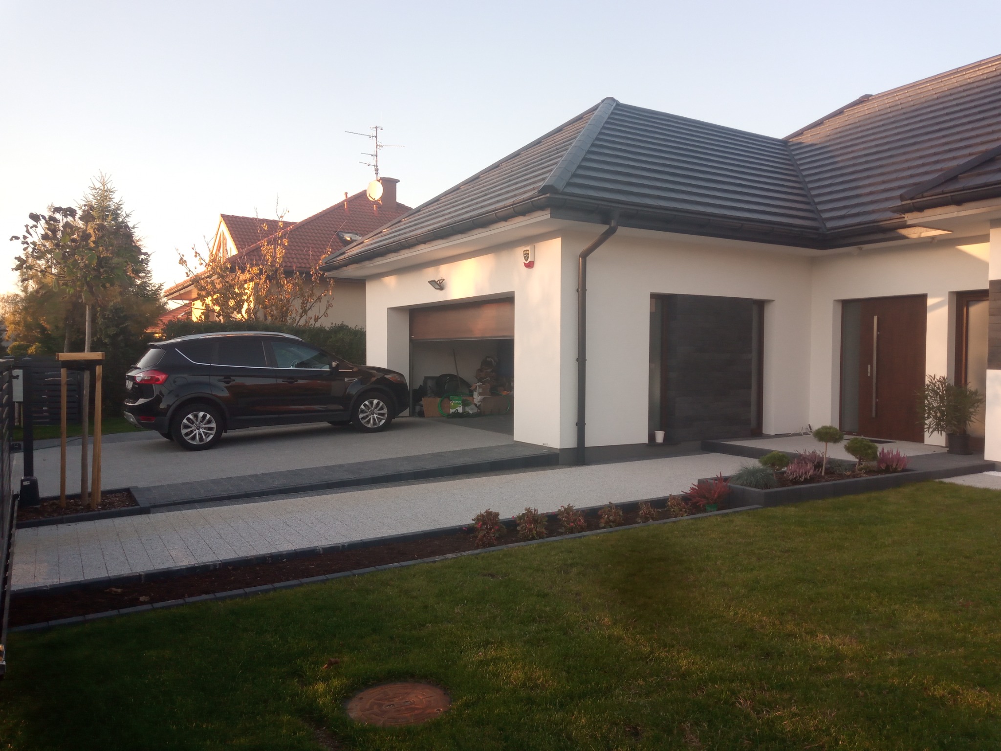 Nowoczesny dom jednorodzinny z szarą kostką brukową na podjeździe i starannie utrzymanym trawnikiem, samochód zaparkowany na podjeździe, garaż z otwartymi drzwiami.