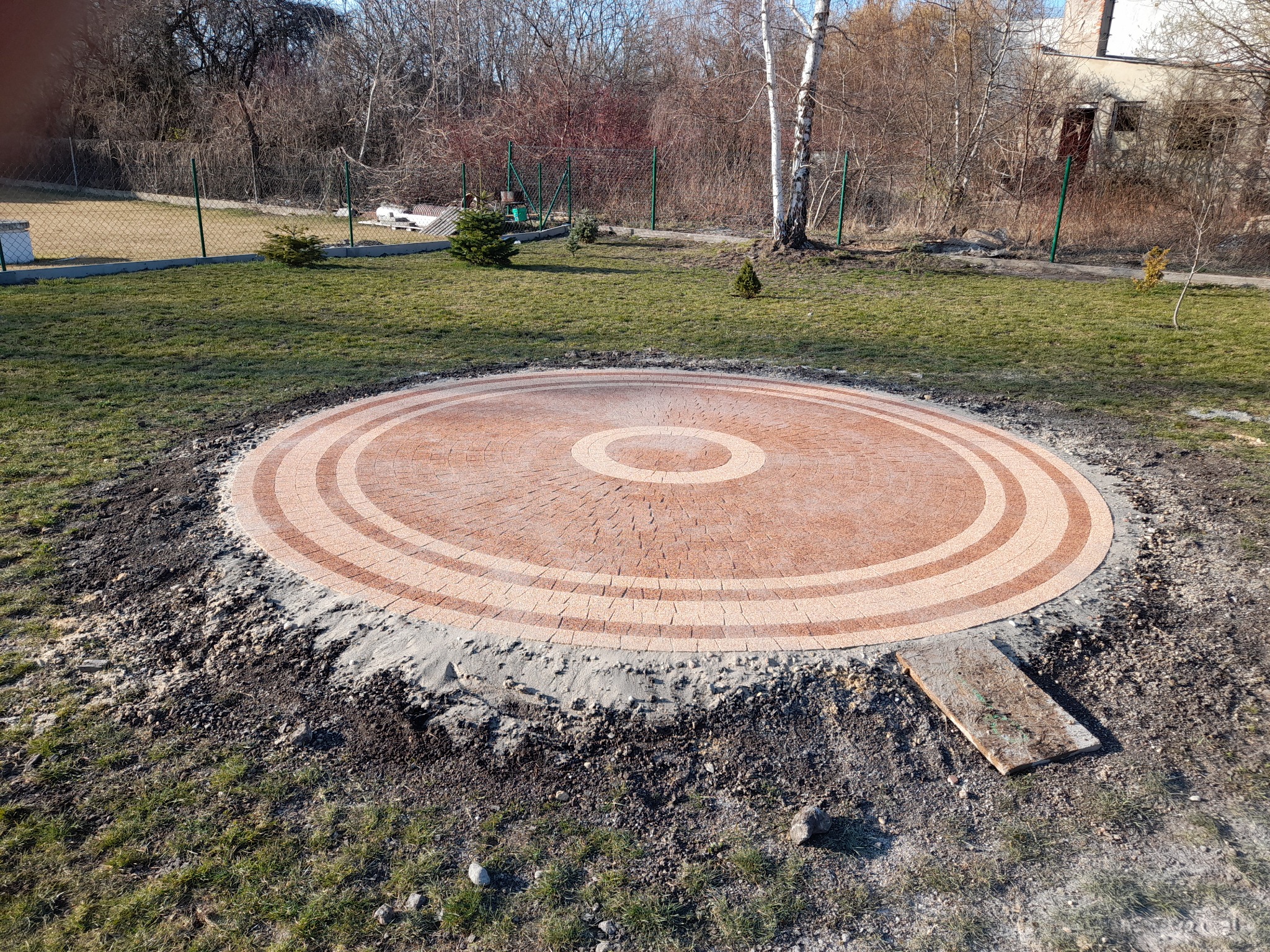 Okrągły plac z kostki brukowej w odcieniach beżu i brązu, otoczony ziemią i trawą, z widoczną deską i młodymi drzewkami w tle.