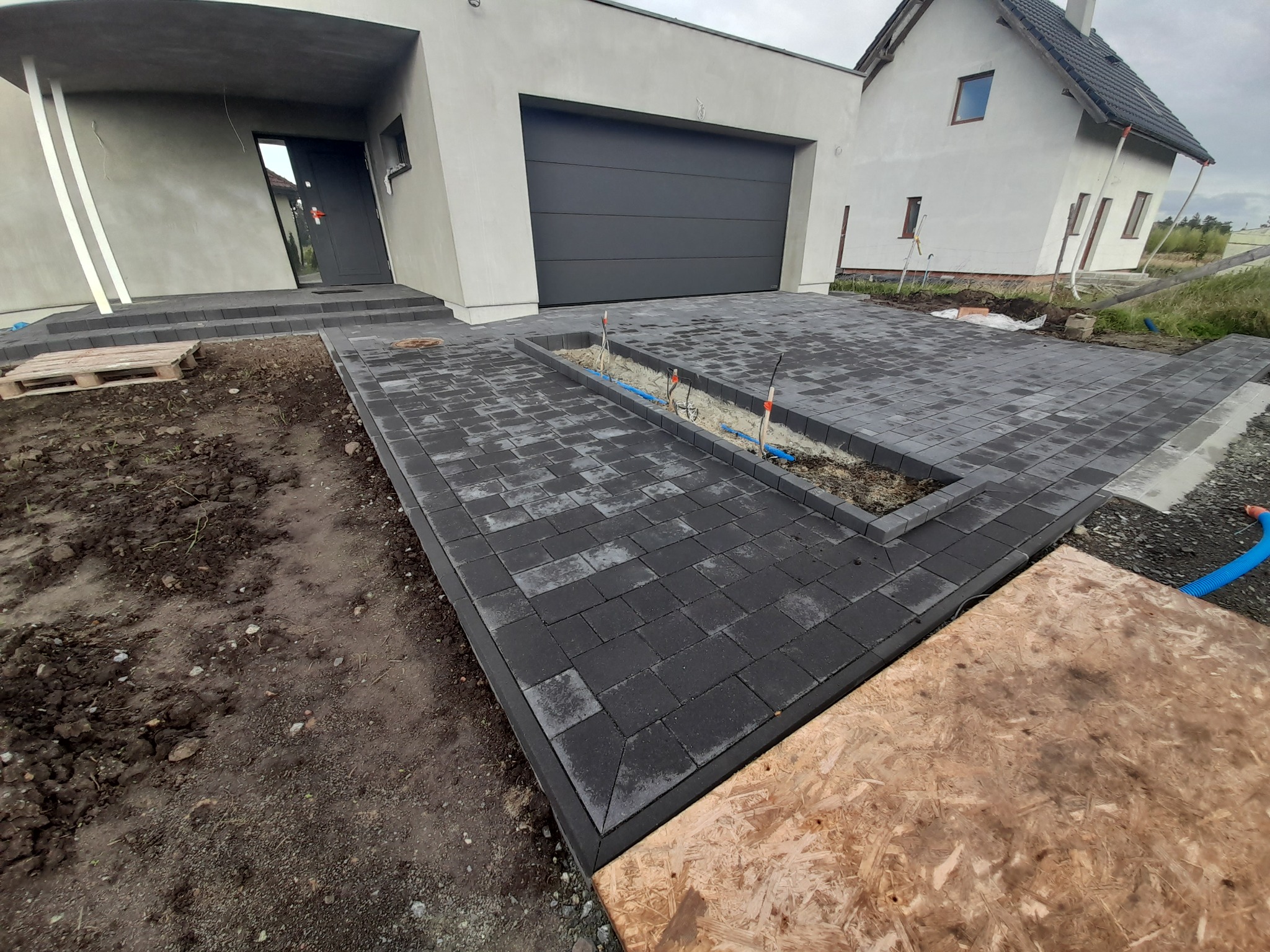 Nowo ułożona, ciemnoszara kostka brukowa przed nowoczesnym domem z garażem, widoczne przygotowane korytko z instalacjami, częściowo zakryte płytą OSB. Na pierwszym planie widoczny fragment palety.