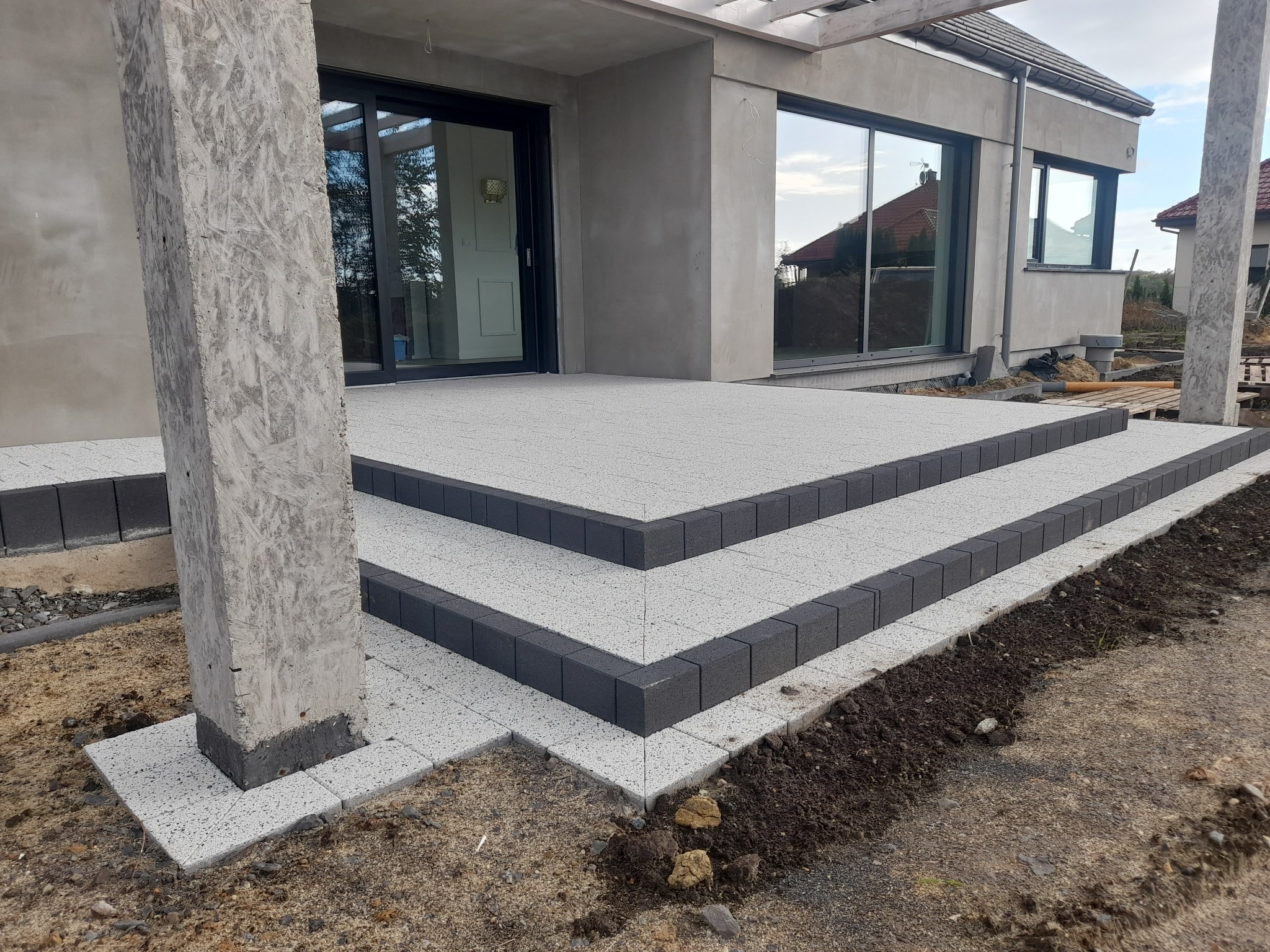 Schody wejściowe do nowoczesnego domu wyłożone jasnoszarą kostką brukową z ciemnoszarym obramowaniem, widoczny surowy betonowy słup podtrzymujący zadaszenie i częściowo wykończona nawierzchnia wokół.